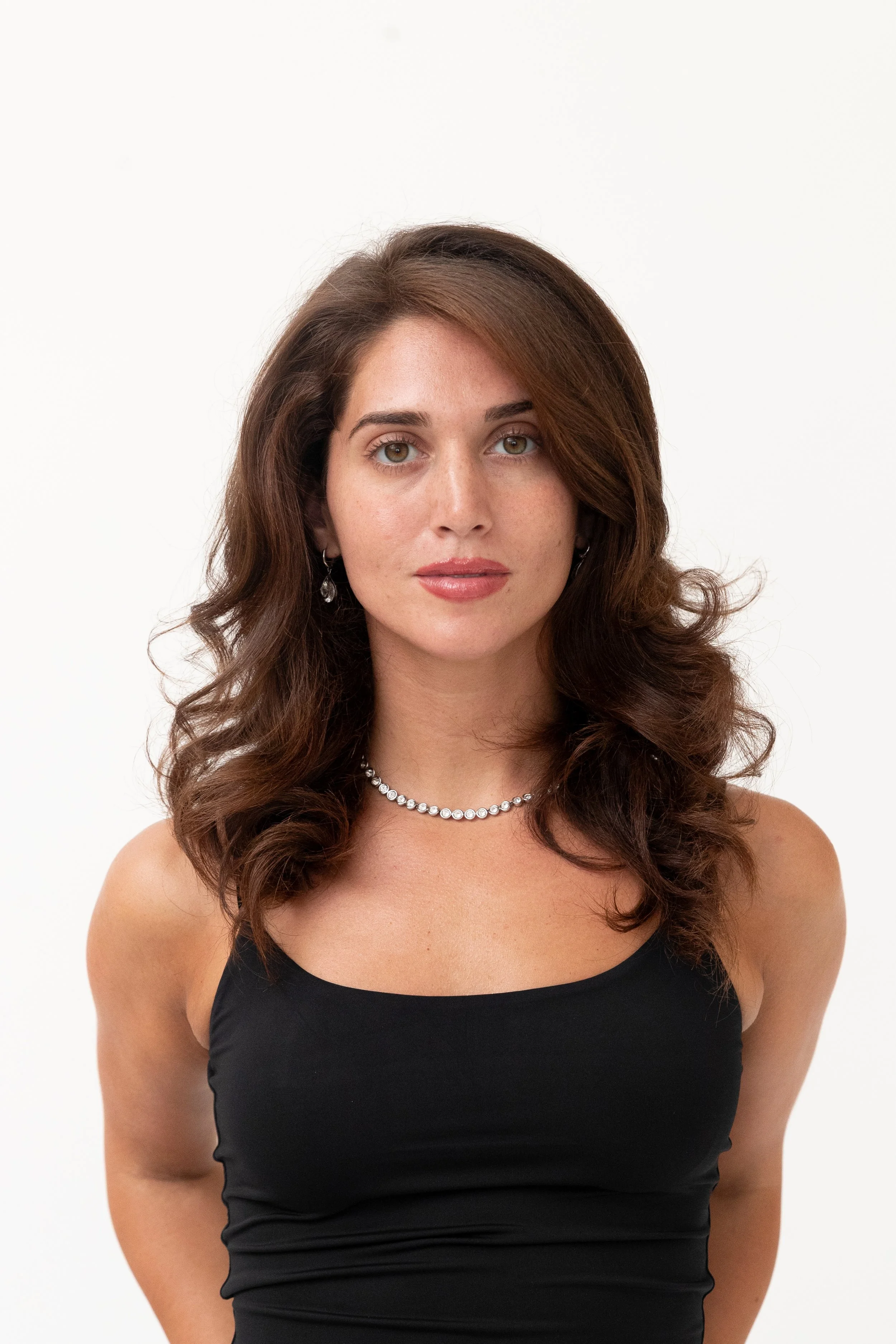 A woman with long, curly brown hair wearing a black top, pearl necklace, and earrings, standing against a plain white background.