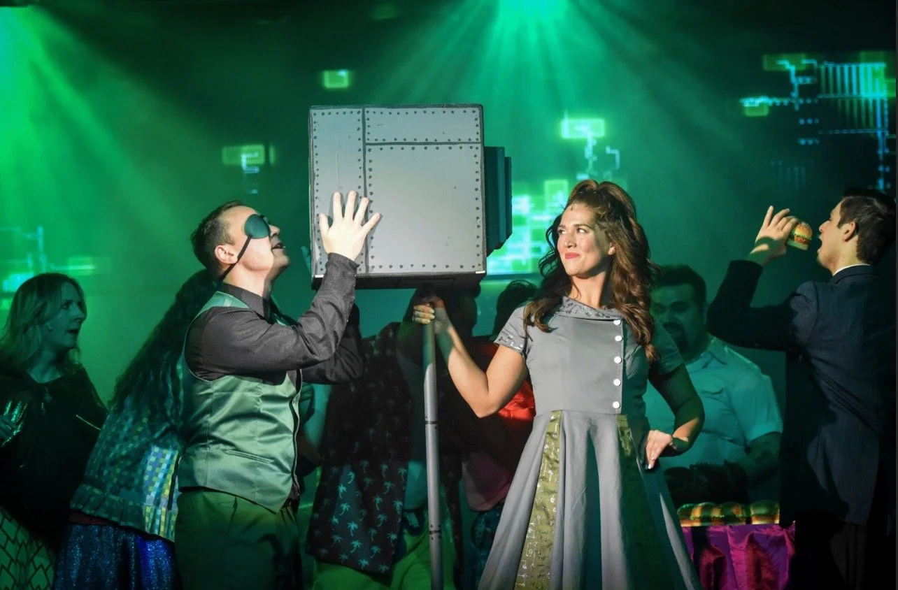 A woman in a vintage style dress holds a large box on a stand, with a person in a headset next to her. Several people are in the background, and green lighting and digital graphics create a futuristic atmosphere.