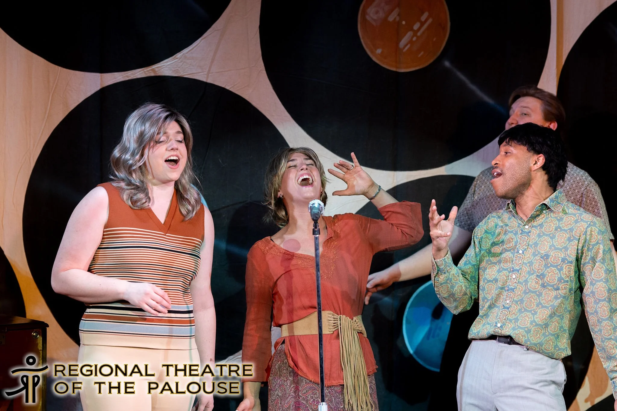 Four people singing on stage with a microphone, set against a colorful abstract backdrop, at the Regional Theatre of the Palouse.