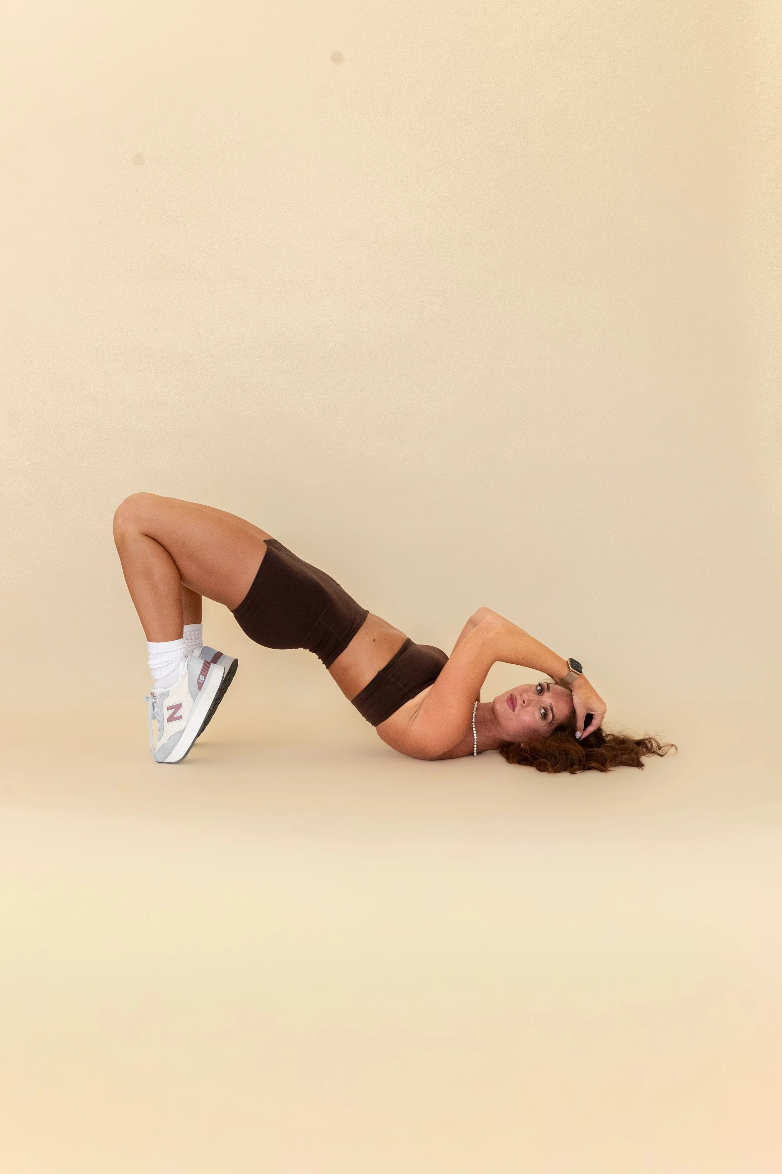 A woman lying on the ground with her back arched, legs bent, and head resting on the floor, wearing athletic shorts, a sports bra, sneakers, white socks, a watch, and a necklace, against a plain beige background.