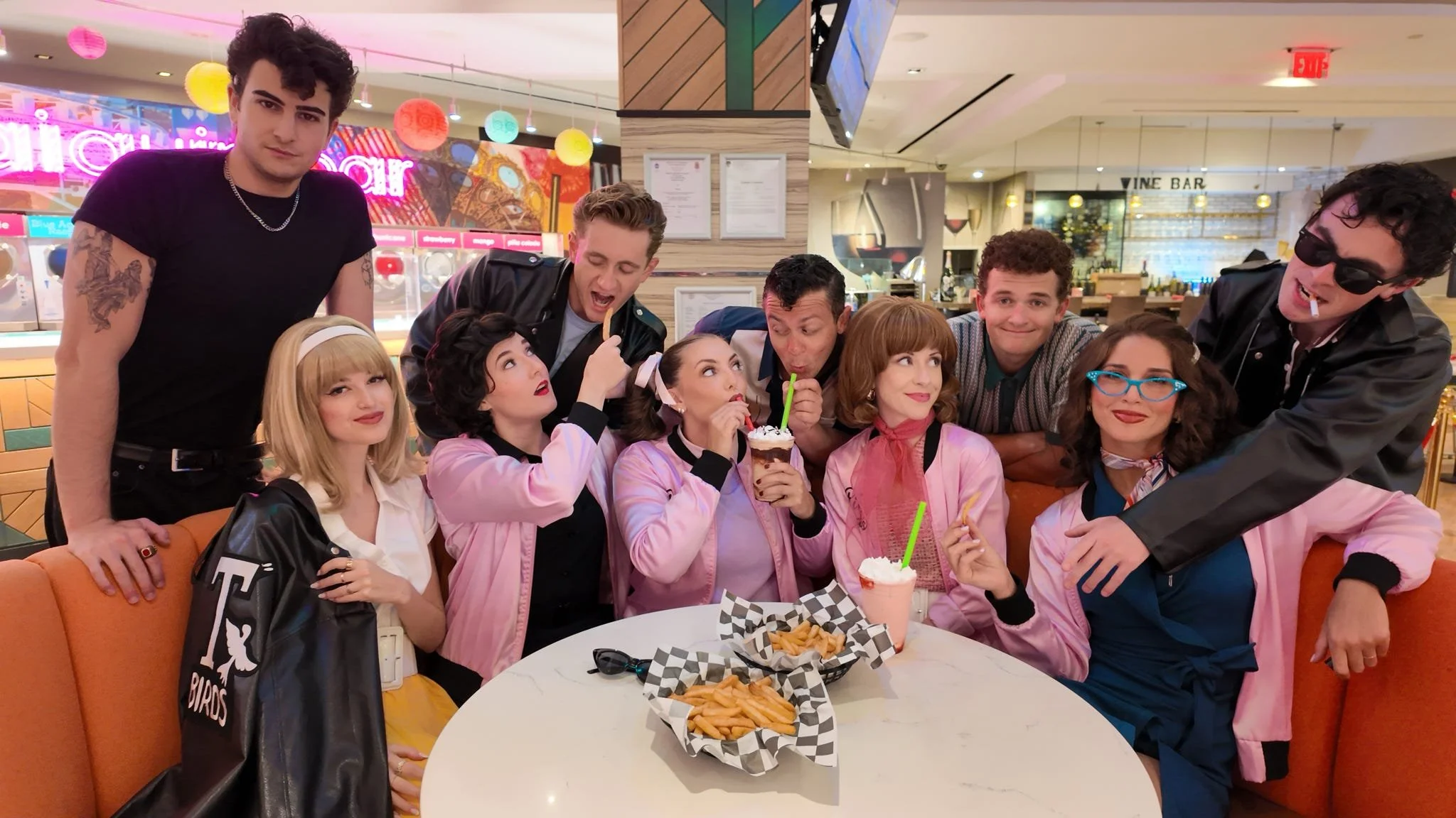 Group of nine young people gathered around a table at a cafe, some wearing pink jackets, enjoying ice cream and snacks, with colorful decor in the background.