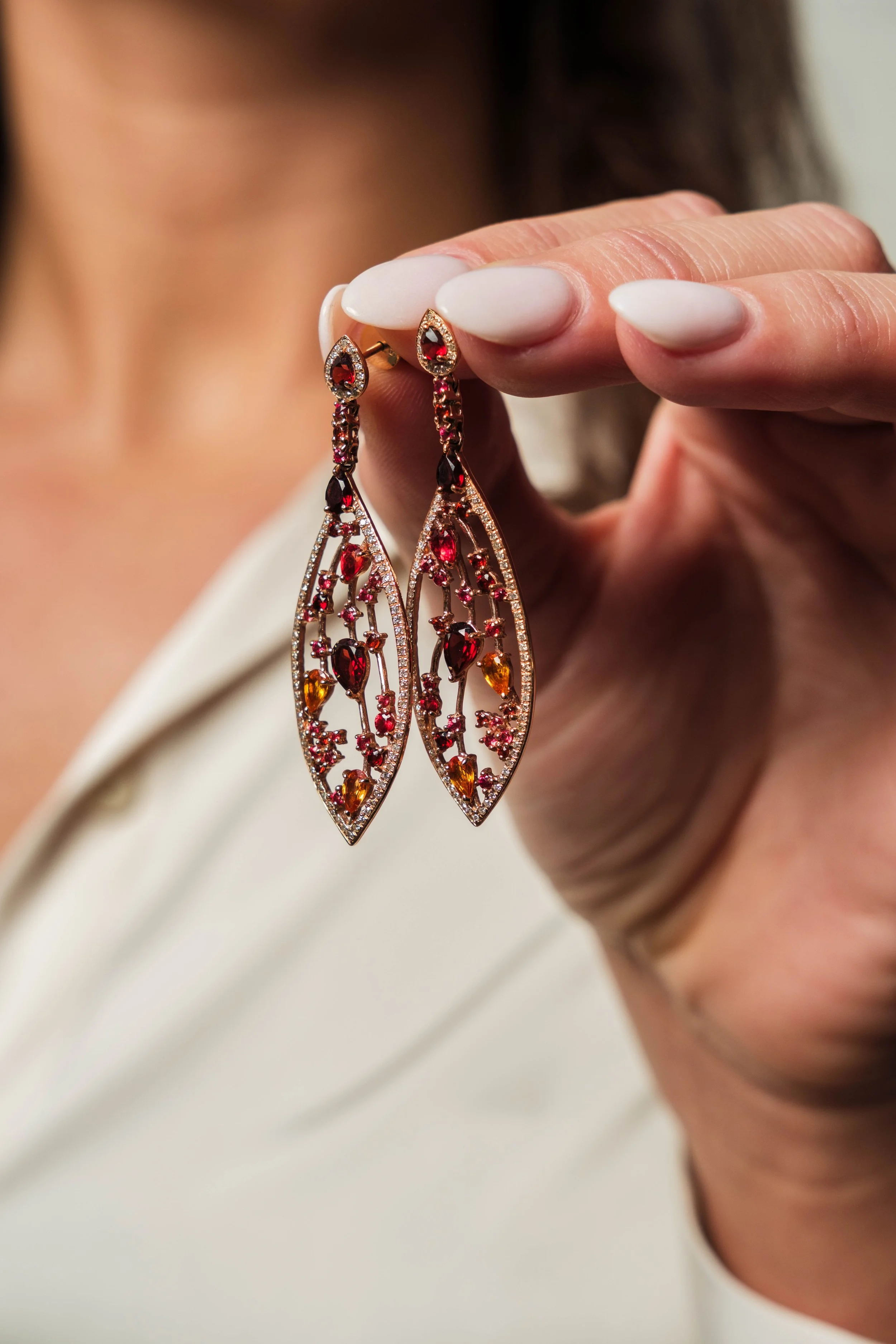 Close-up of a person's hand holding a pair of ornate chandelier earrings with red and orange gemstones.