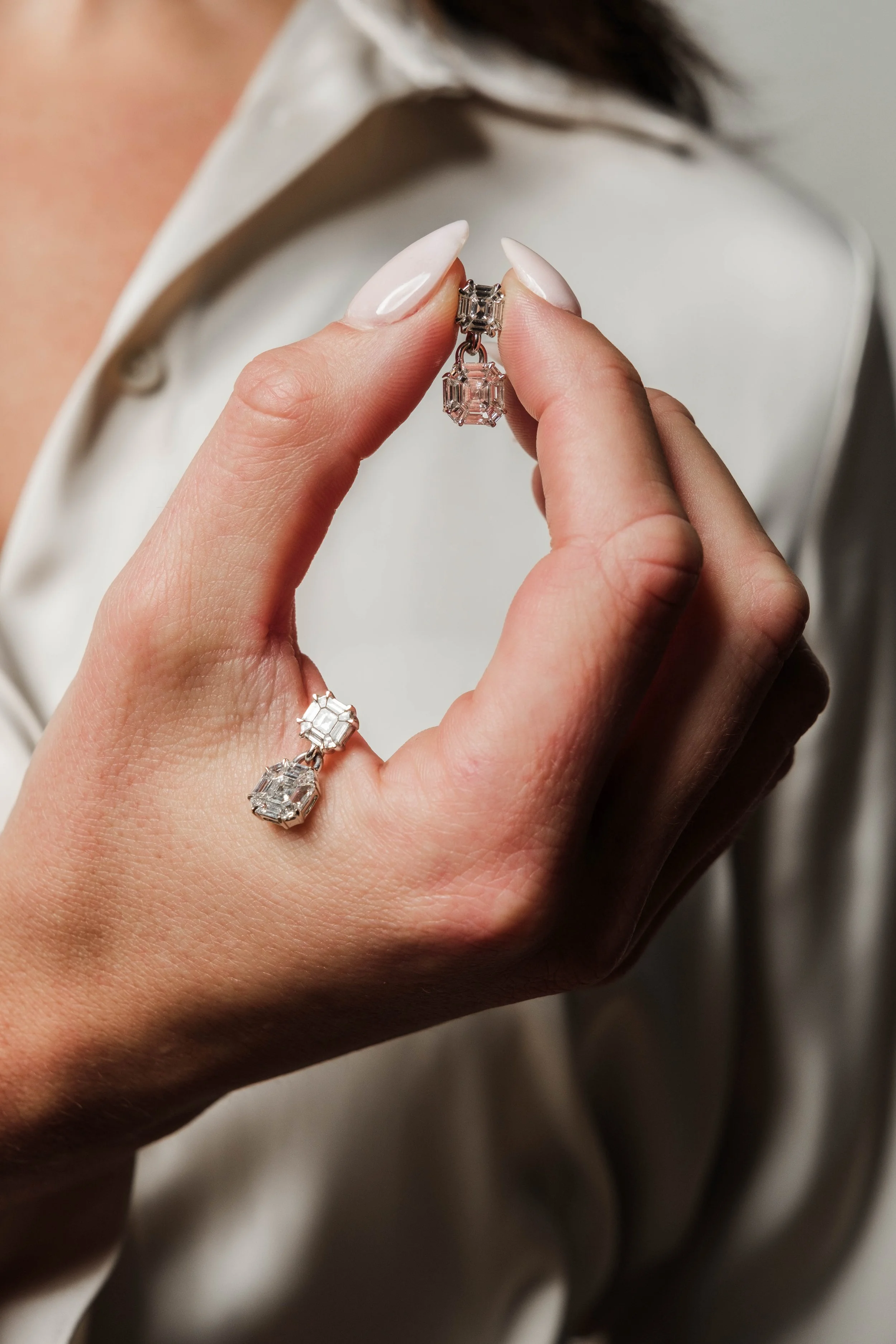 A person's hand holding a pair of diamond earrings in front of their white shirt and dark hair.