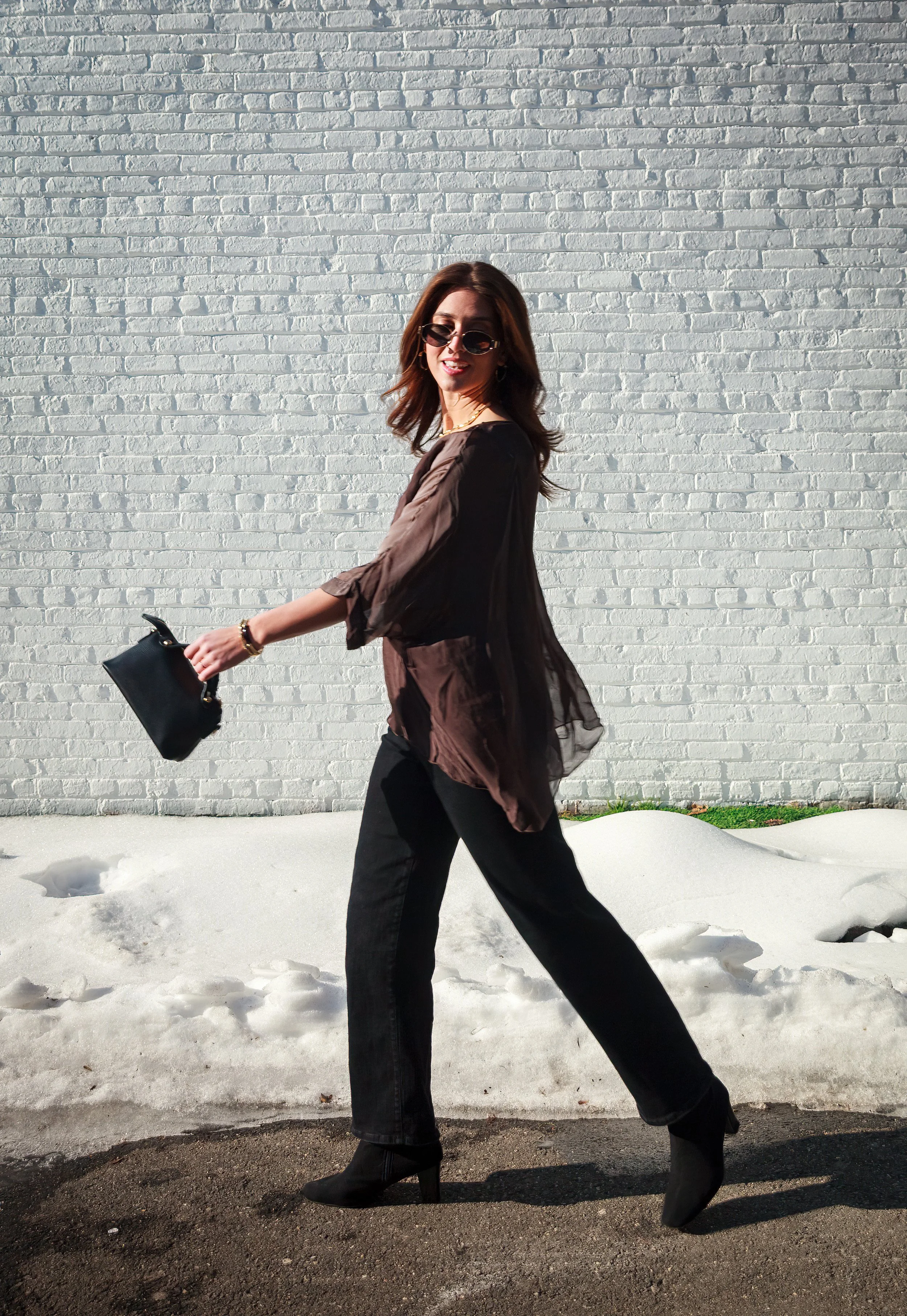A woman with brown hair, wearing sunglasses, a sheer brown cover-up, black pants, and high-heeled boots, walking outdoors on snow, holding a black handbag against a white brick wall.