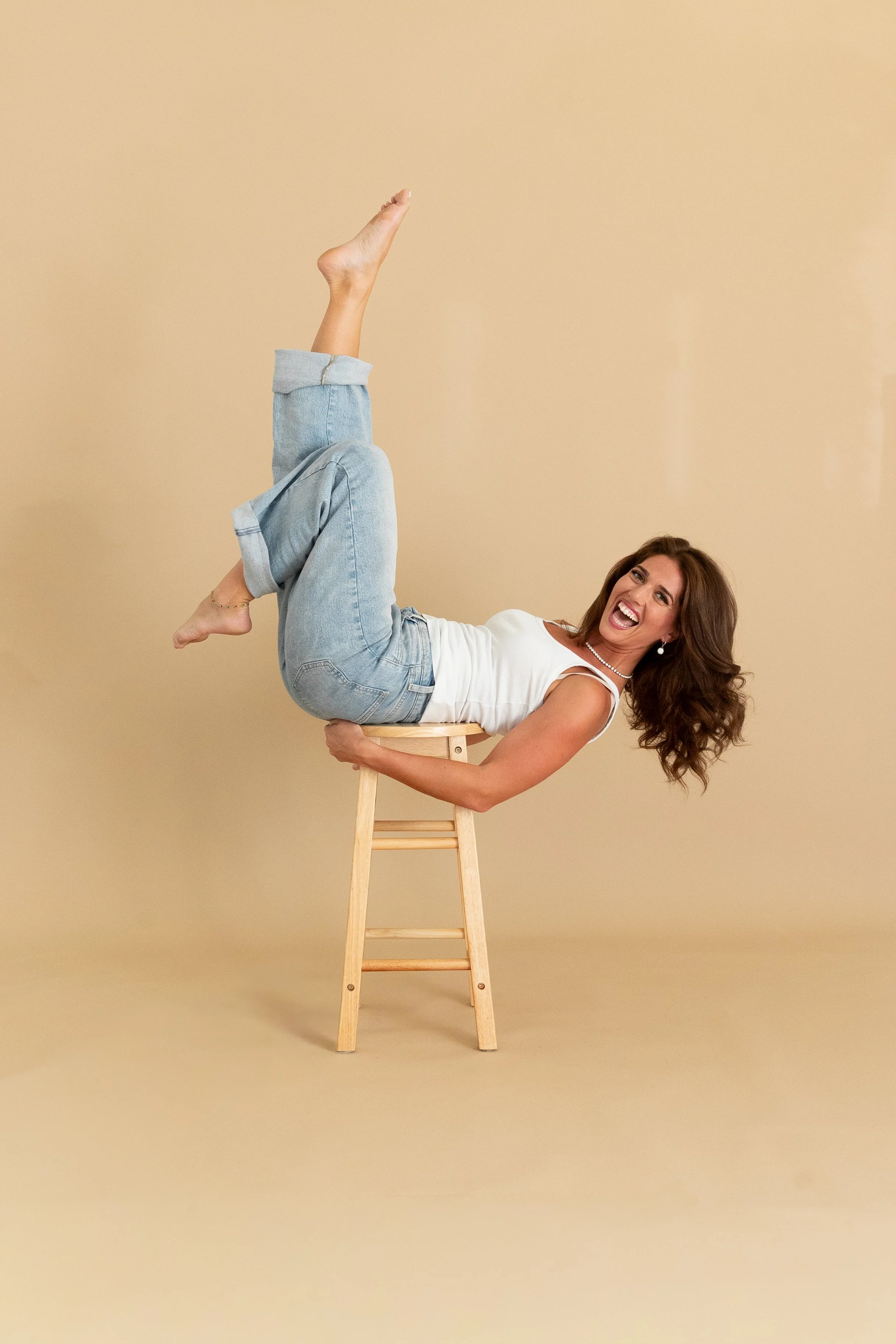 A woman with long brown hair, wearing a white sleeveless top and blue jeans, is lying on a wooden stool with her body upside down, one leg raised, smiling happily, against a neutral beige background.