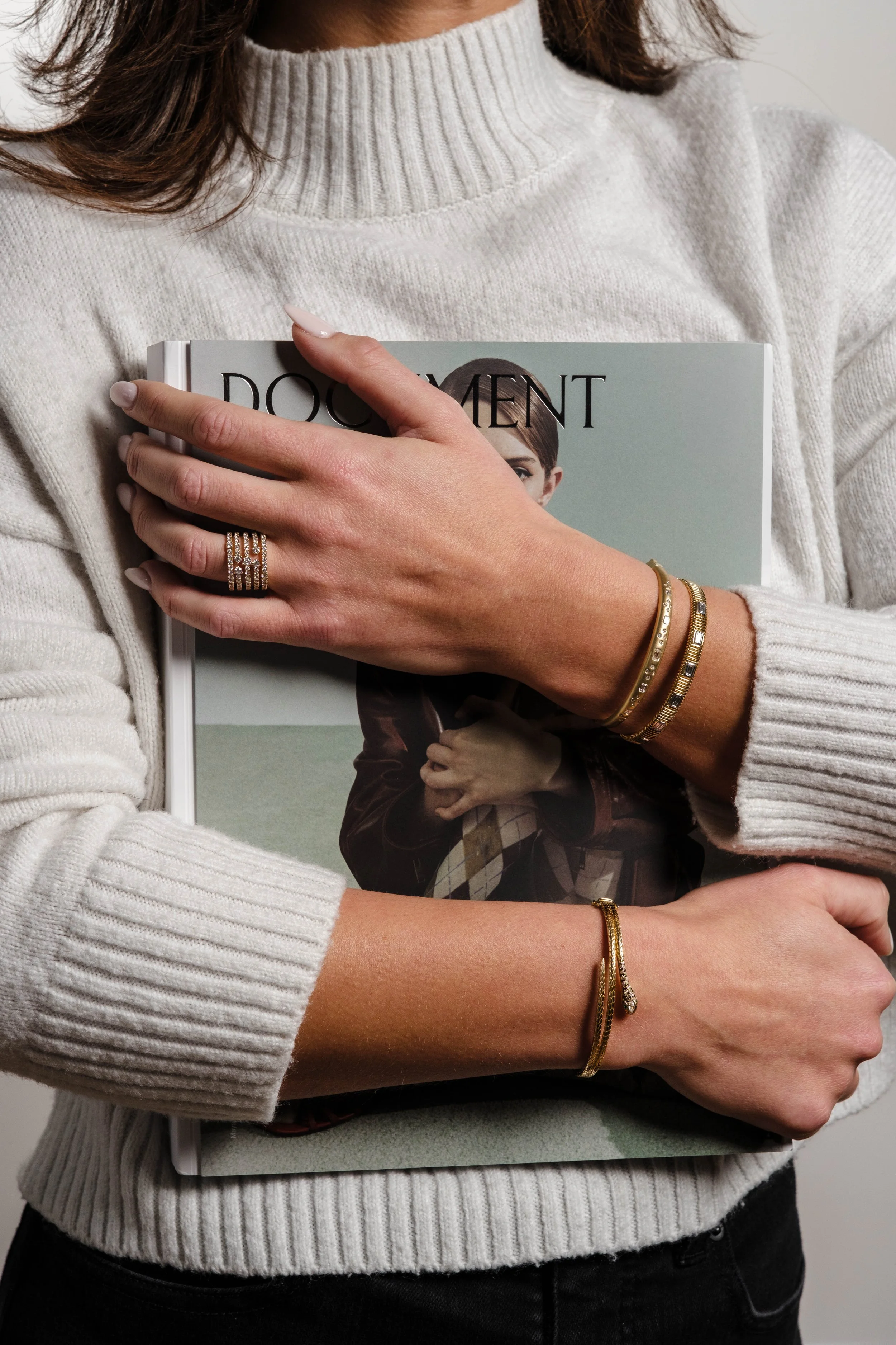 Woman in a cream-colored turtleneck sweater holding a magazine titled "Document" with her right hand, wearing gold rings and bracelets.
