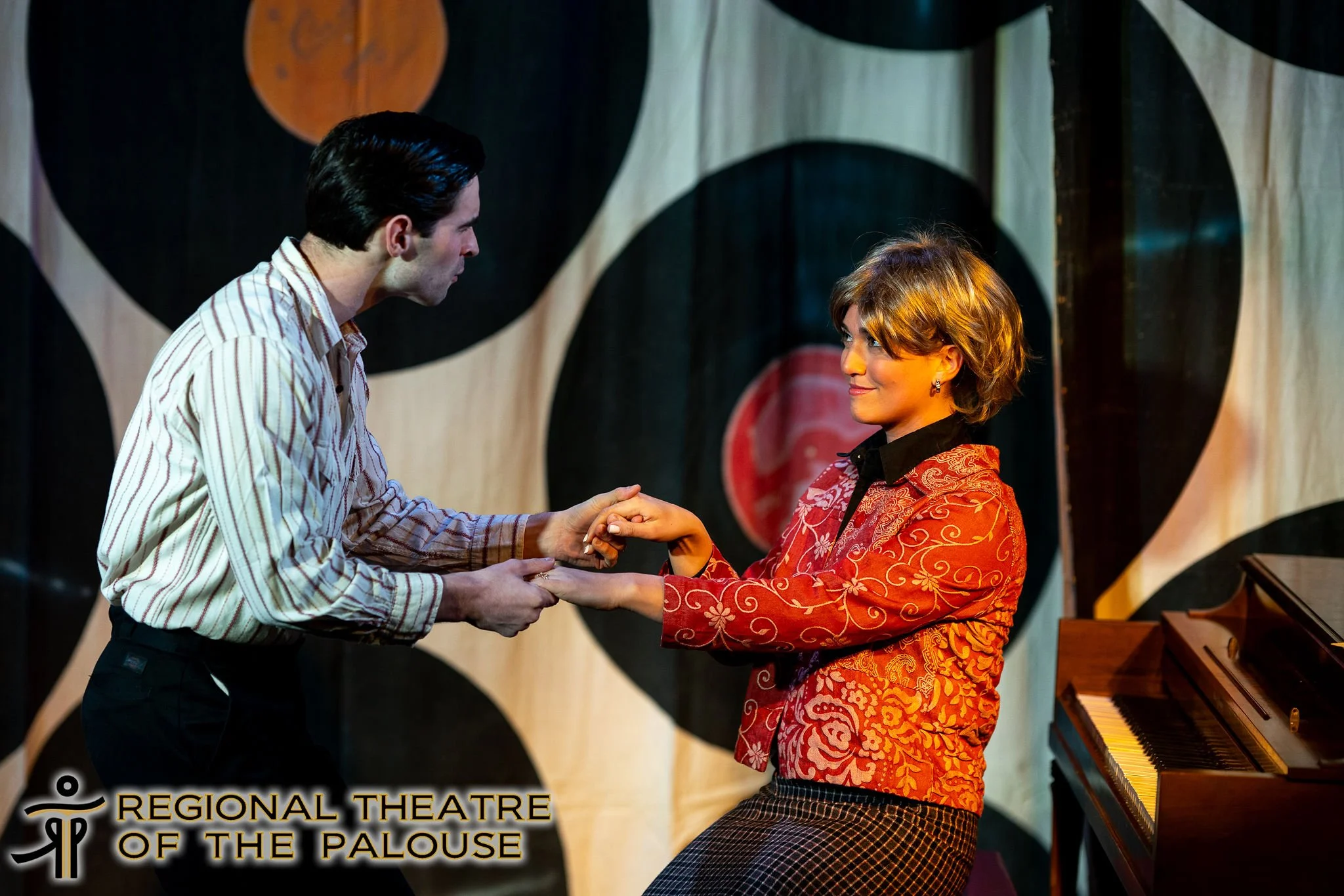 A man and a woman are holding hands on stage, facing each other. The woman is wearing a red jacket with floral patterns and the man is wearing a striped shirt. They are in a theater setting with a colorful abstract backdrop and a piano nearby.