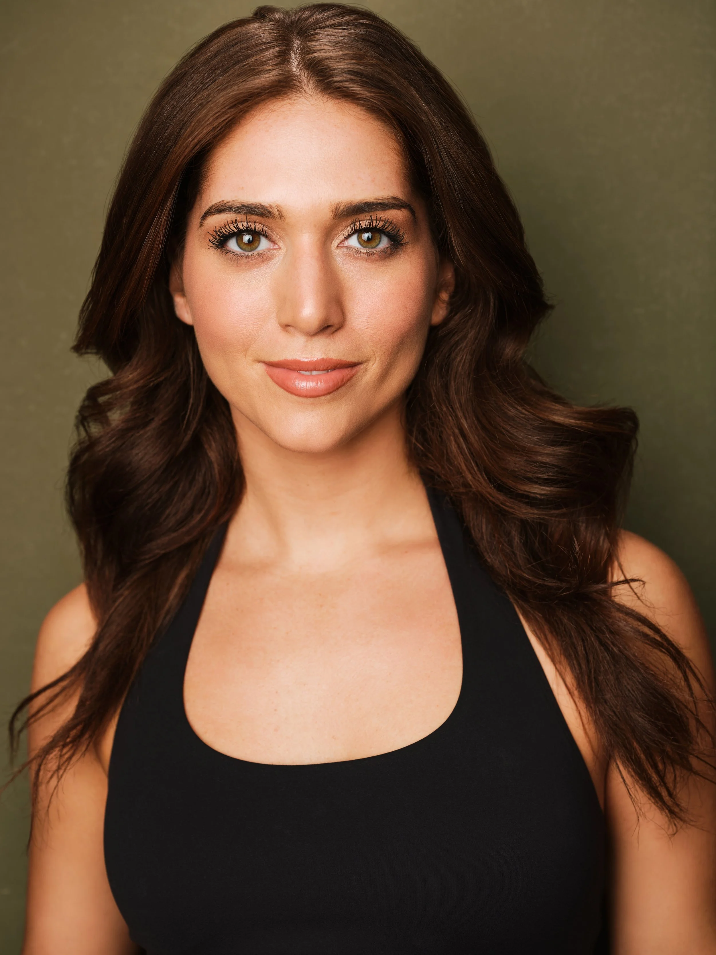 Close-up portrait of a woman with wavy brown hair, hazel eyes, wearing a black top, smiling softly against a plain olive green background.