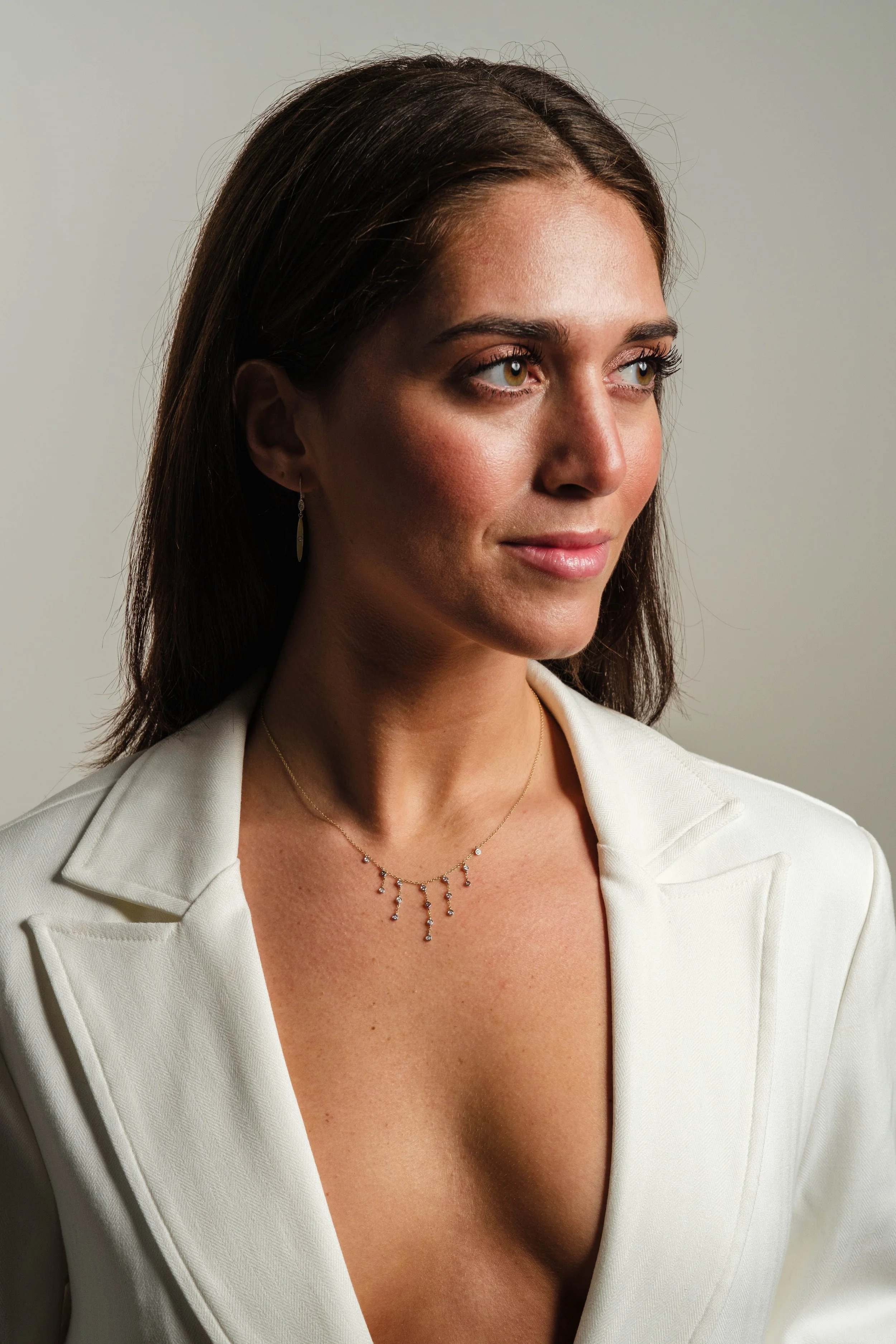 A woman with light brown hair, wearing a white blazer, a delicate necklace, and drop earrings, looking slightly to the right with a soft smile.