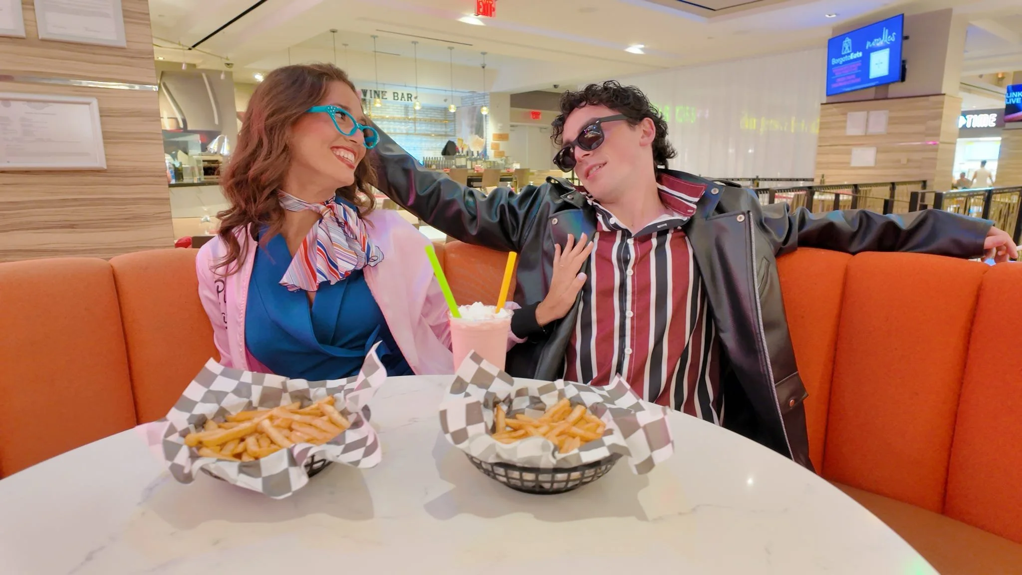 Two people enjoying a meal at a restaurant, smiling and wearing sunglasses. There are baskets of French fries, a milkshake, and a plate of food on the table.