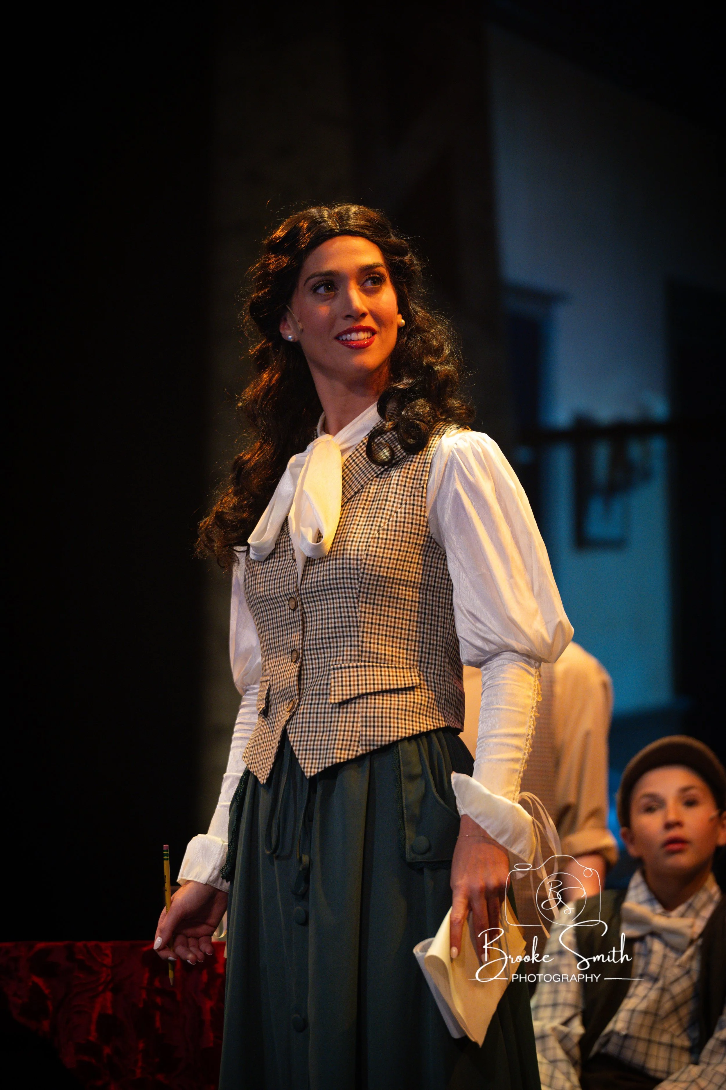 A woman with dark, curly hair, wearing period costume clothing, stands on stage with others in background. She is smiling and holding a paper and pencil.