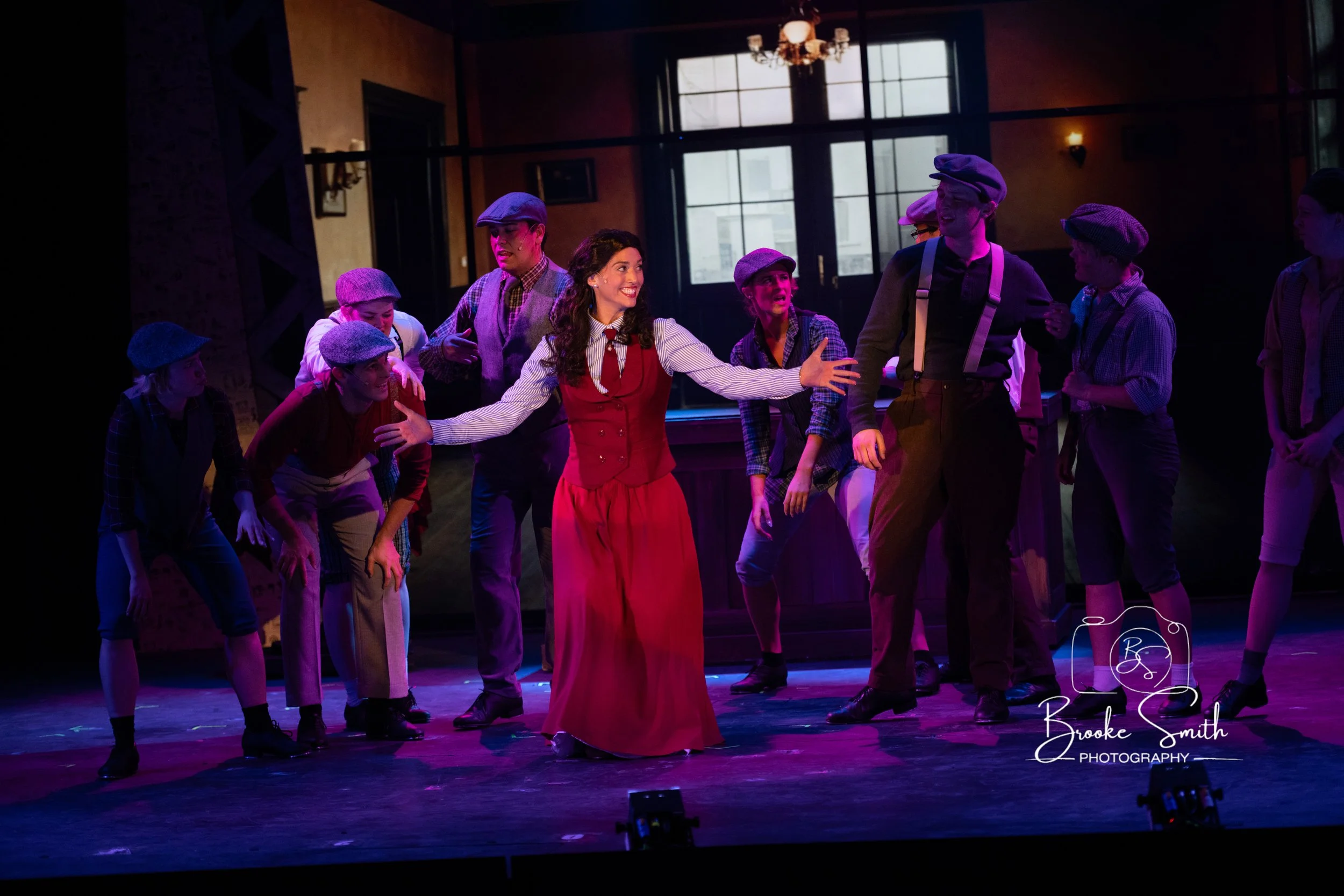 A group of performers on stage, including a woman in a red dress with arms outstretched, surrounded by children dressed in vintage clothing and berets, with a spotlight highlighting their expressions and movements.