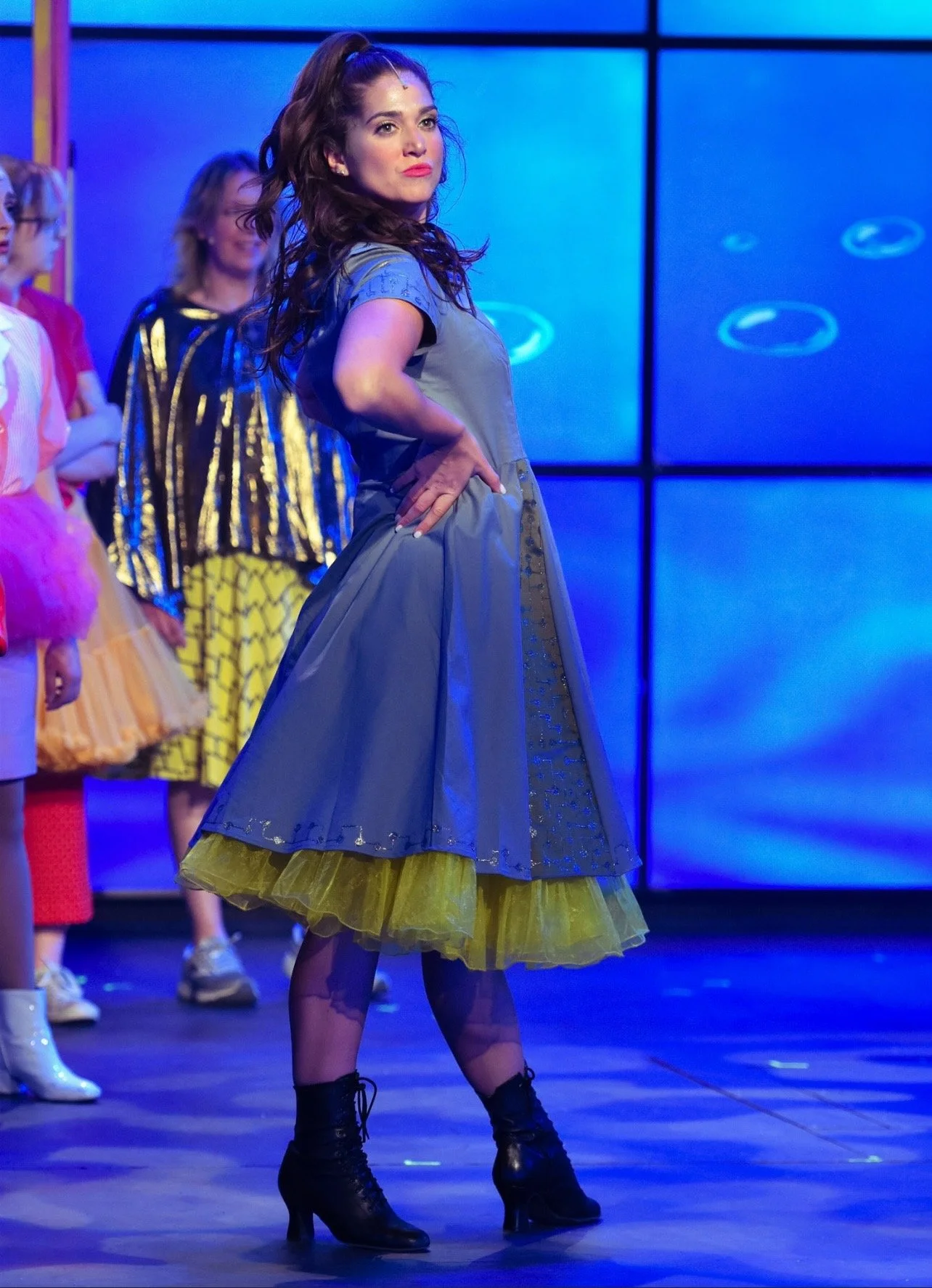 A woman in a colorful, layered dress with a gray top and yellow tutu-like skirt, wearing black lace-up boots, striking a confident pose on stage. Behind her, other women in vibrant outfits are visible, suggesting a theatrical or dance performance.