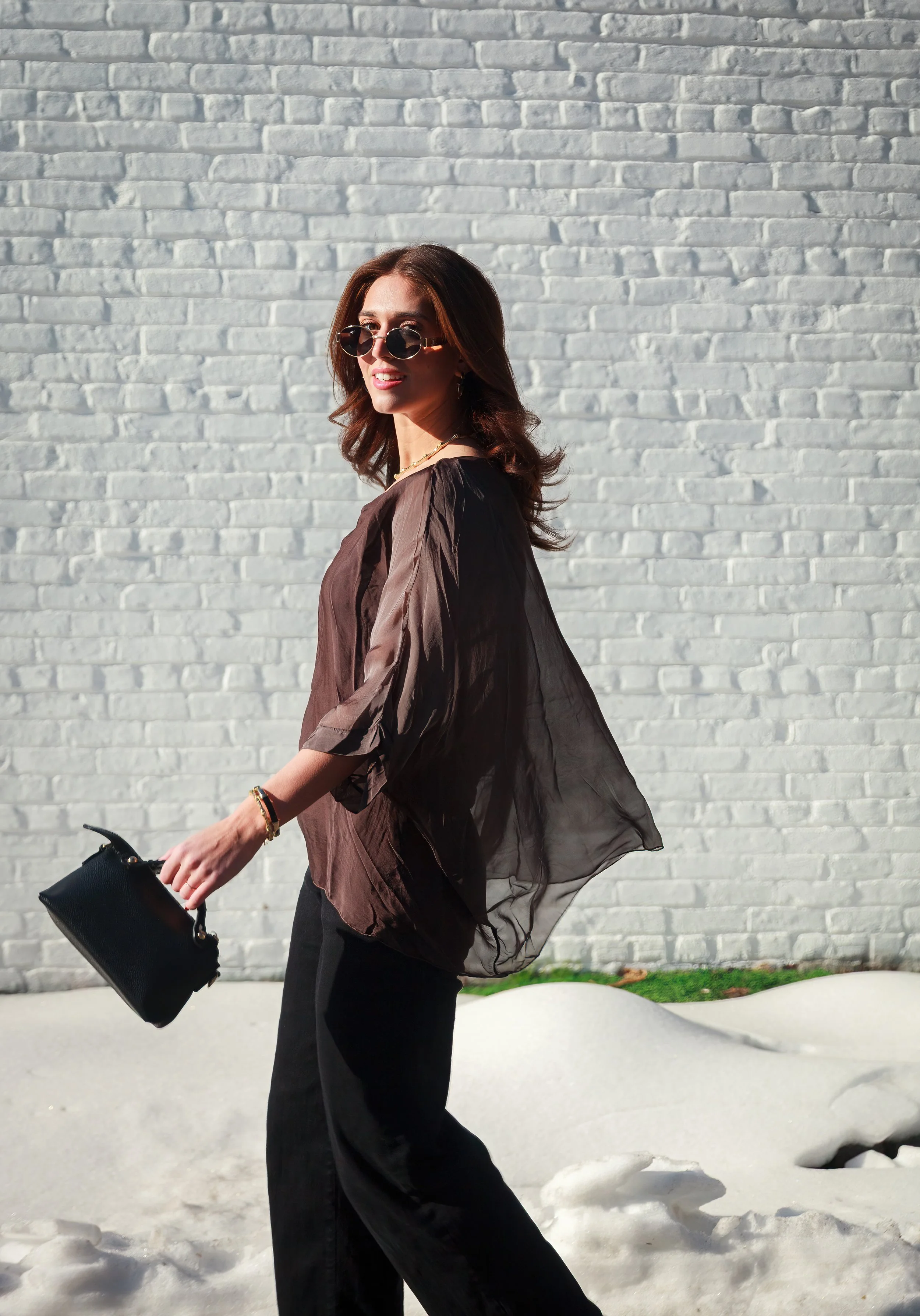 A woman with brown hair and sunglasses walking outdoors on a sunny day, holding a black handbag, with a white brick wall and some snow on the ground in the background.