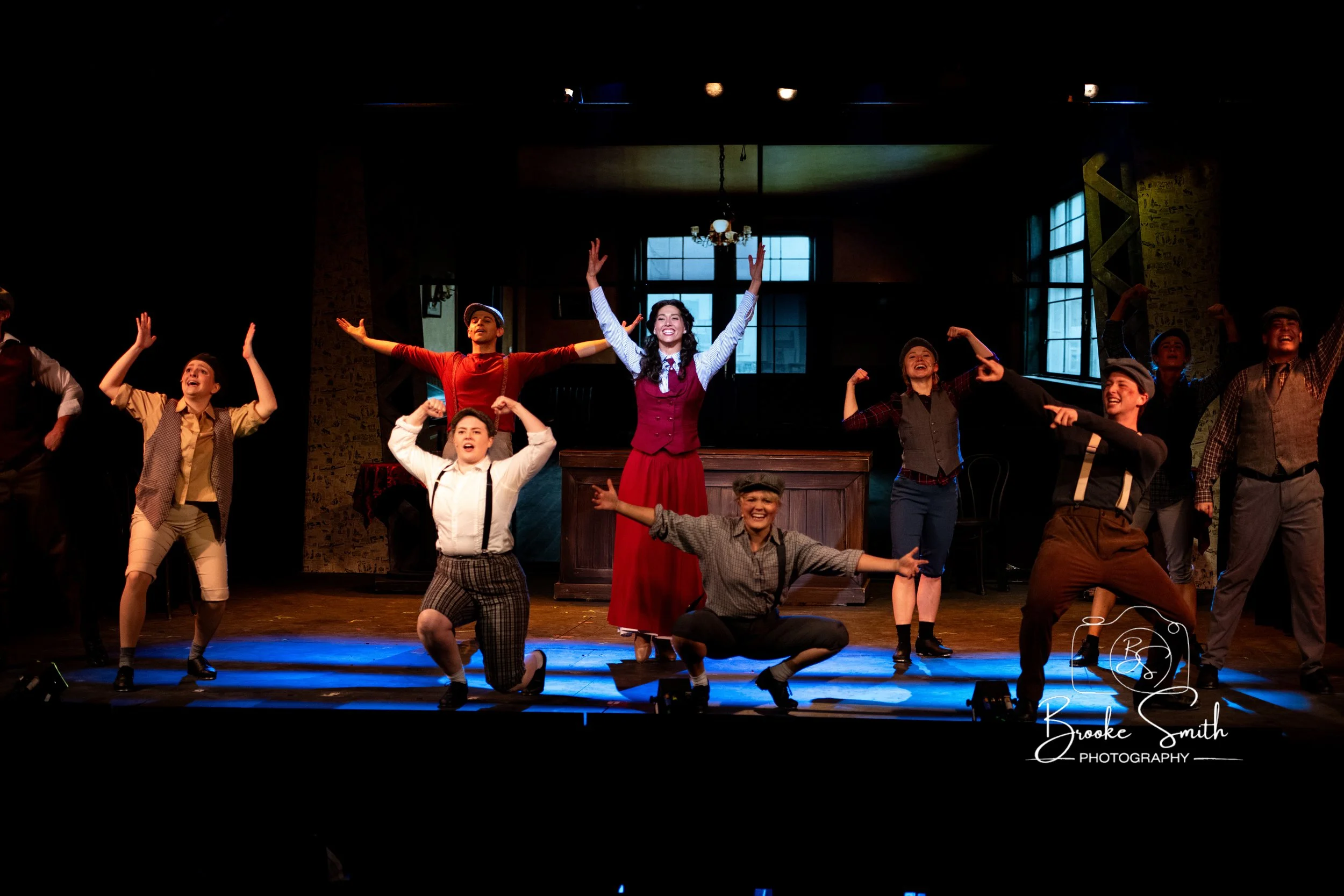 Actors performing on stage in a theatrical production with diverse costumes, with a woman in the center raising her arms and smiling, surrounded by other performers in various poses and expressions.