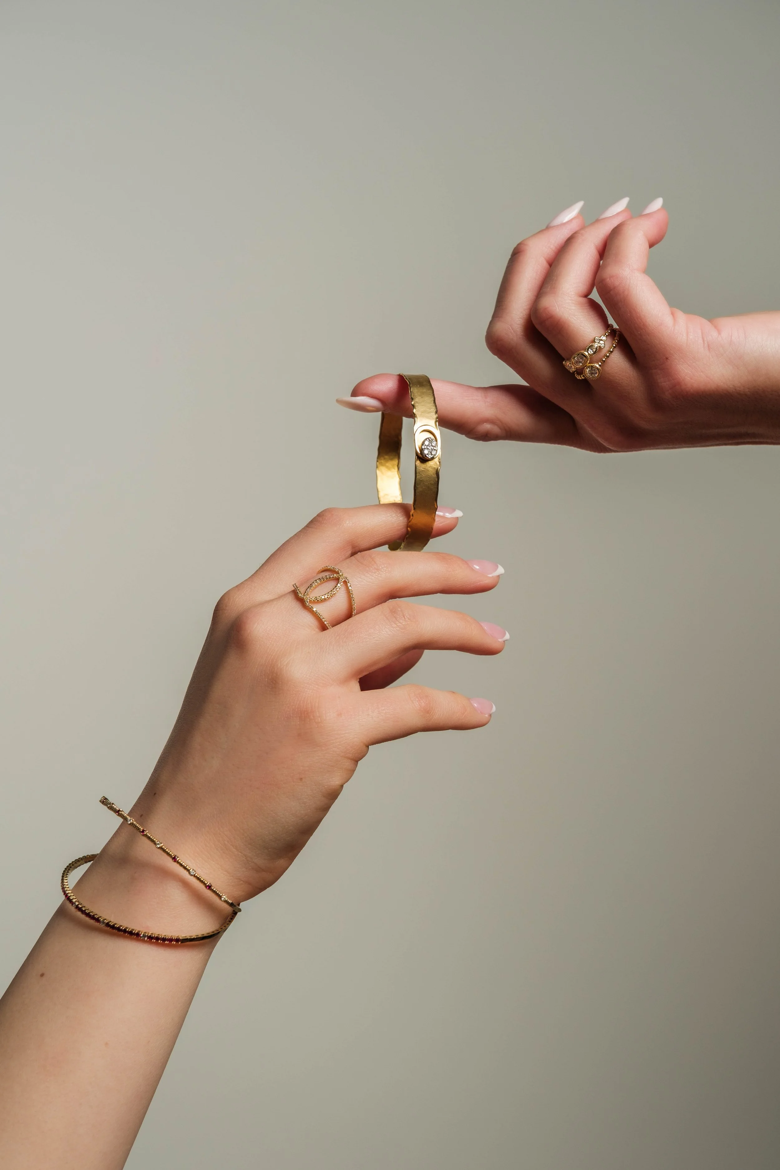 Two hands with light skin tone holding jewelry, including a gold bracelet and rings, with a plain beige background.