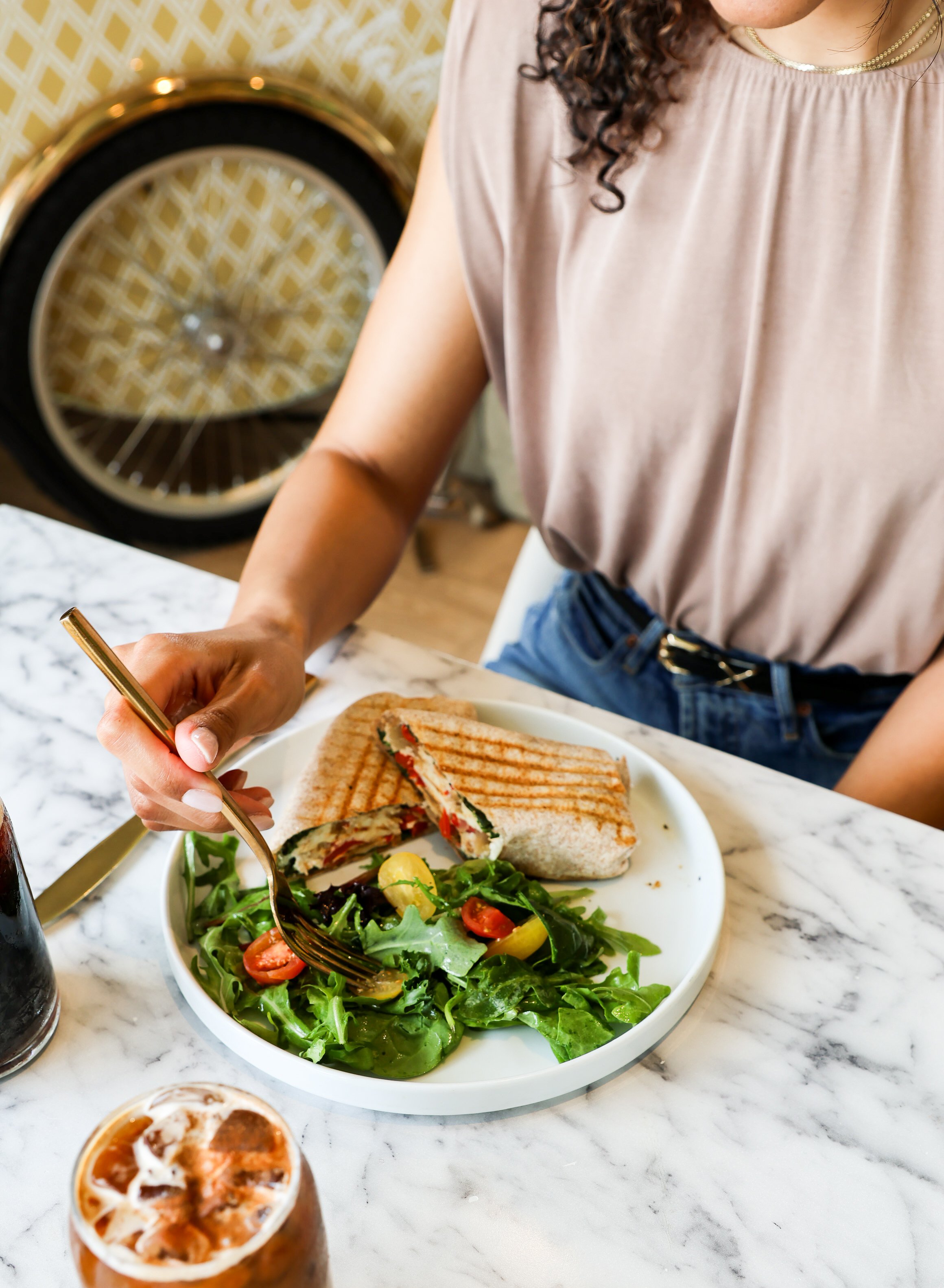 Partially visible woman with curly hair wearing a beige top and necklaces, sitting at a marble table with a plate of grilled sandwich, salad, and two cold drinks.