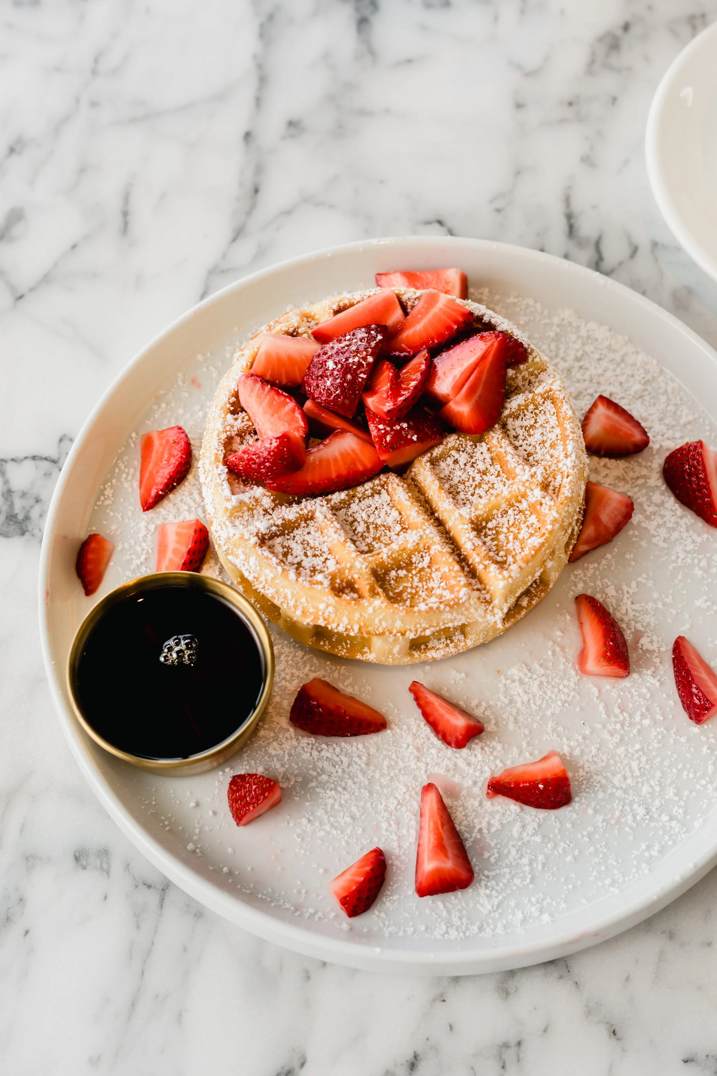 A waffle topped with sliced strawberries and powdered sugar, served on a white plate with a side of syrup in a small container, on a marble surface.