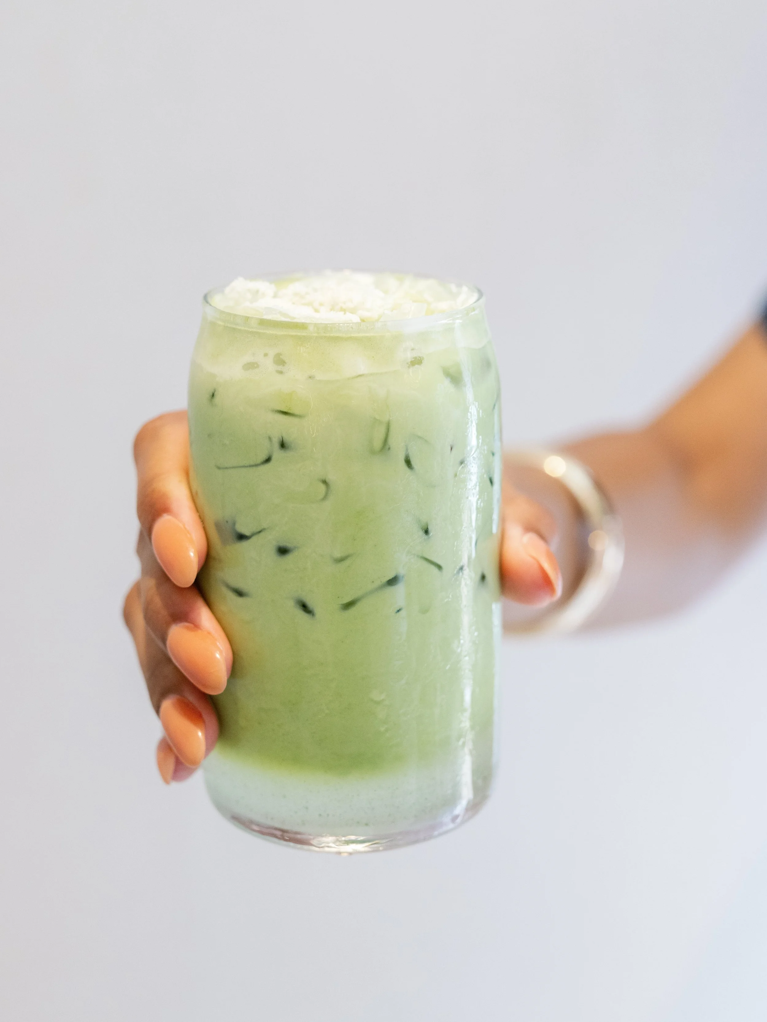 A person's hand holding a glass of iced matcha latte topped with whipped cream against a plain white background.