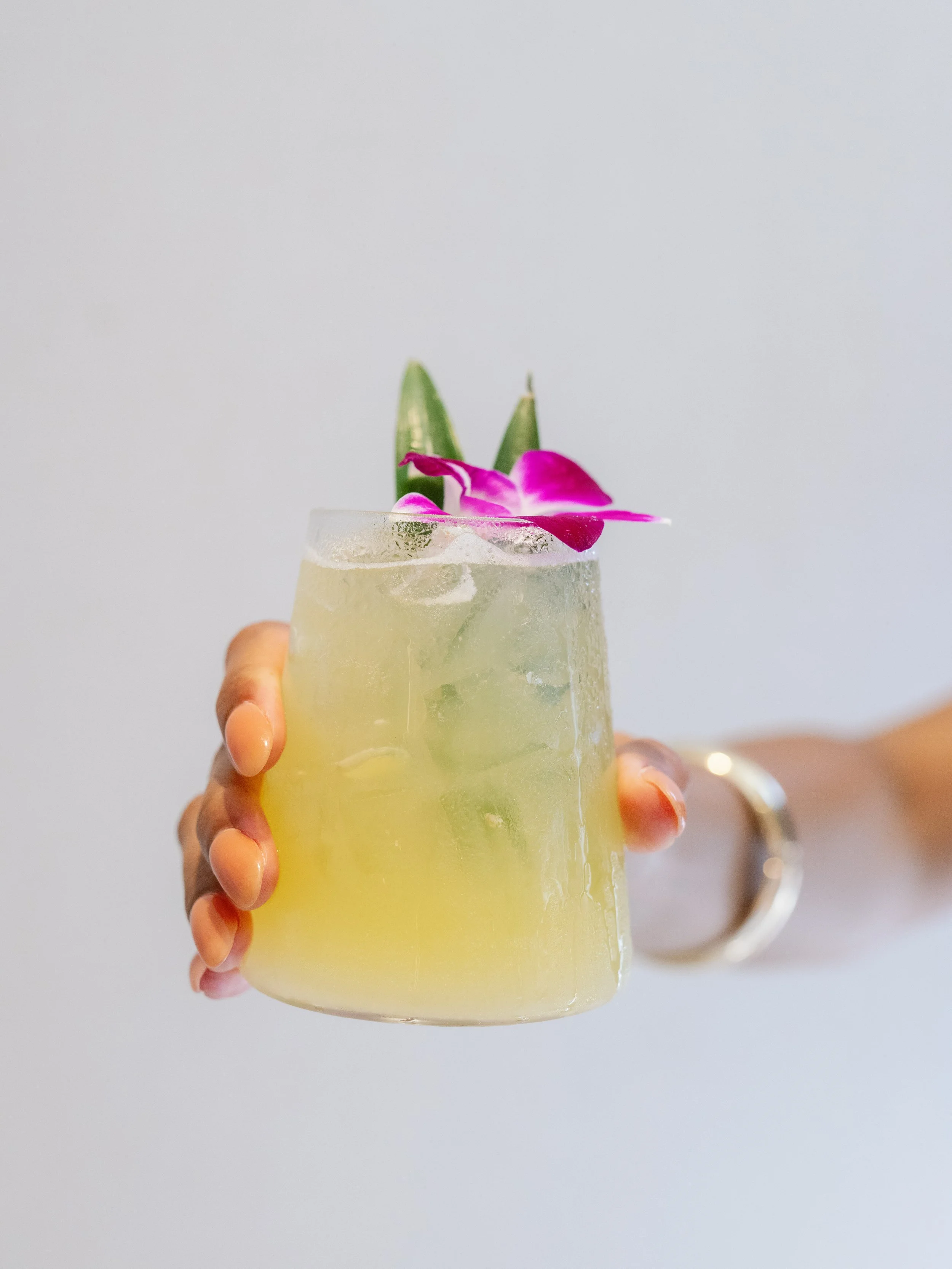 A hand holding a tall glass of light yellow cocktail with ice, garnished with purple orchid and green leaves, against a plain white background.