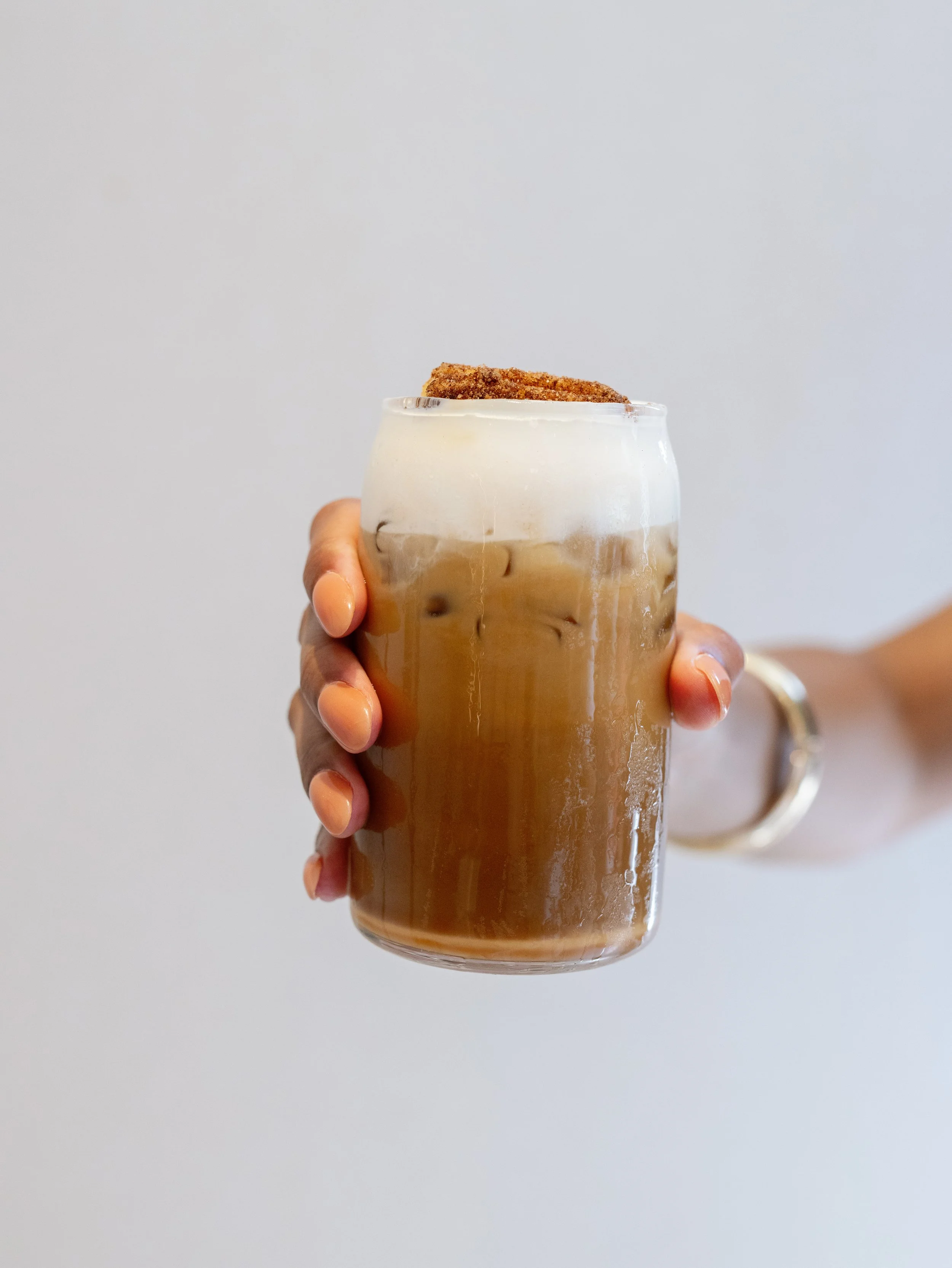 Person holding a glass of iced coffee topped with whipped cream and a cinnamon stick against a plain light background.