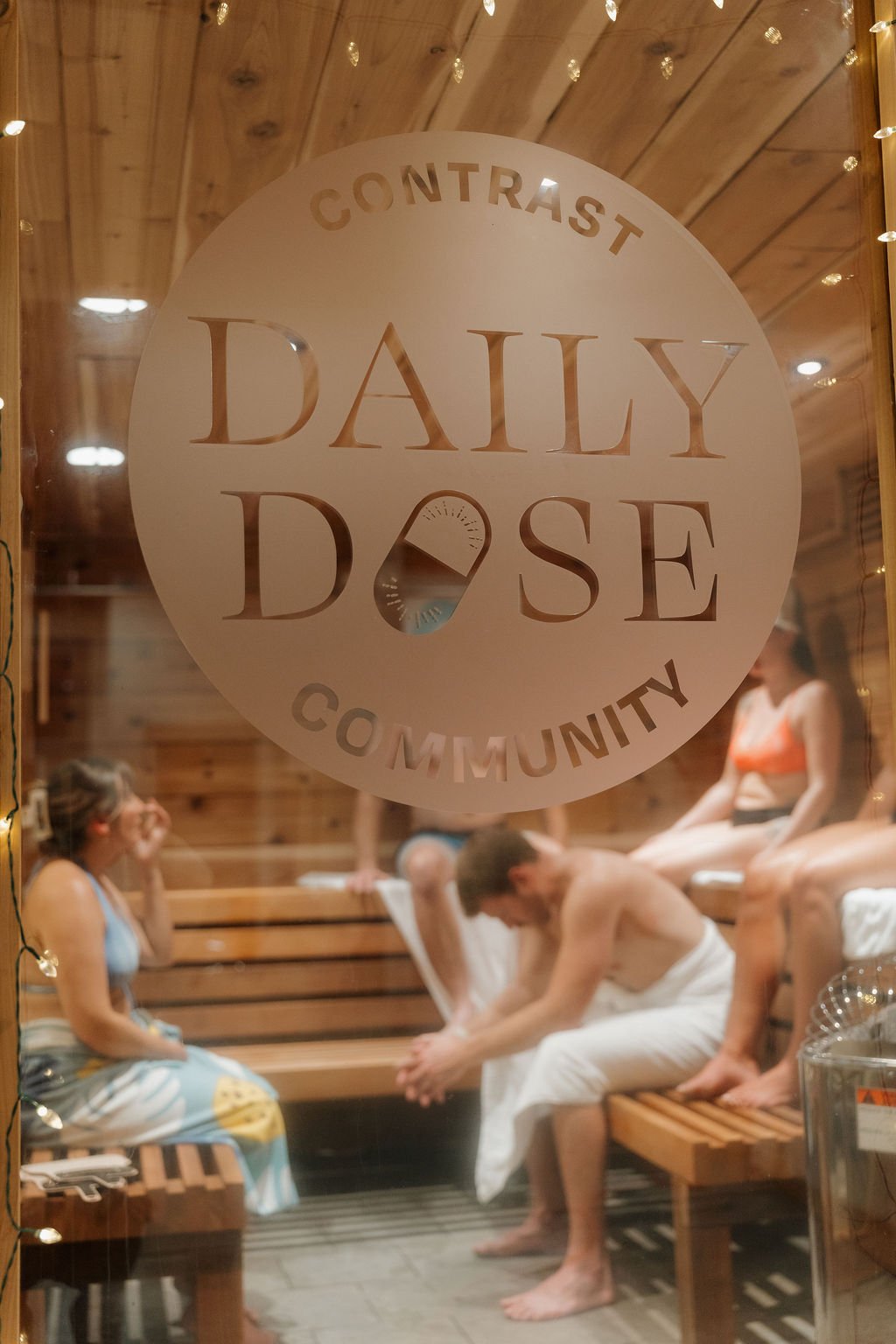A woman with wet hair and skin, wearing a colorful bikini top and a gold necklace, standing with her eyes closed in a wooden sauna or steam room.