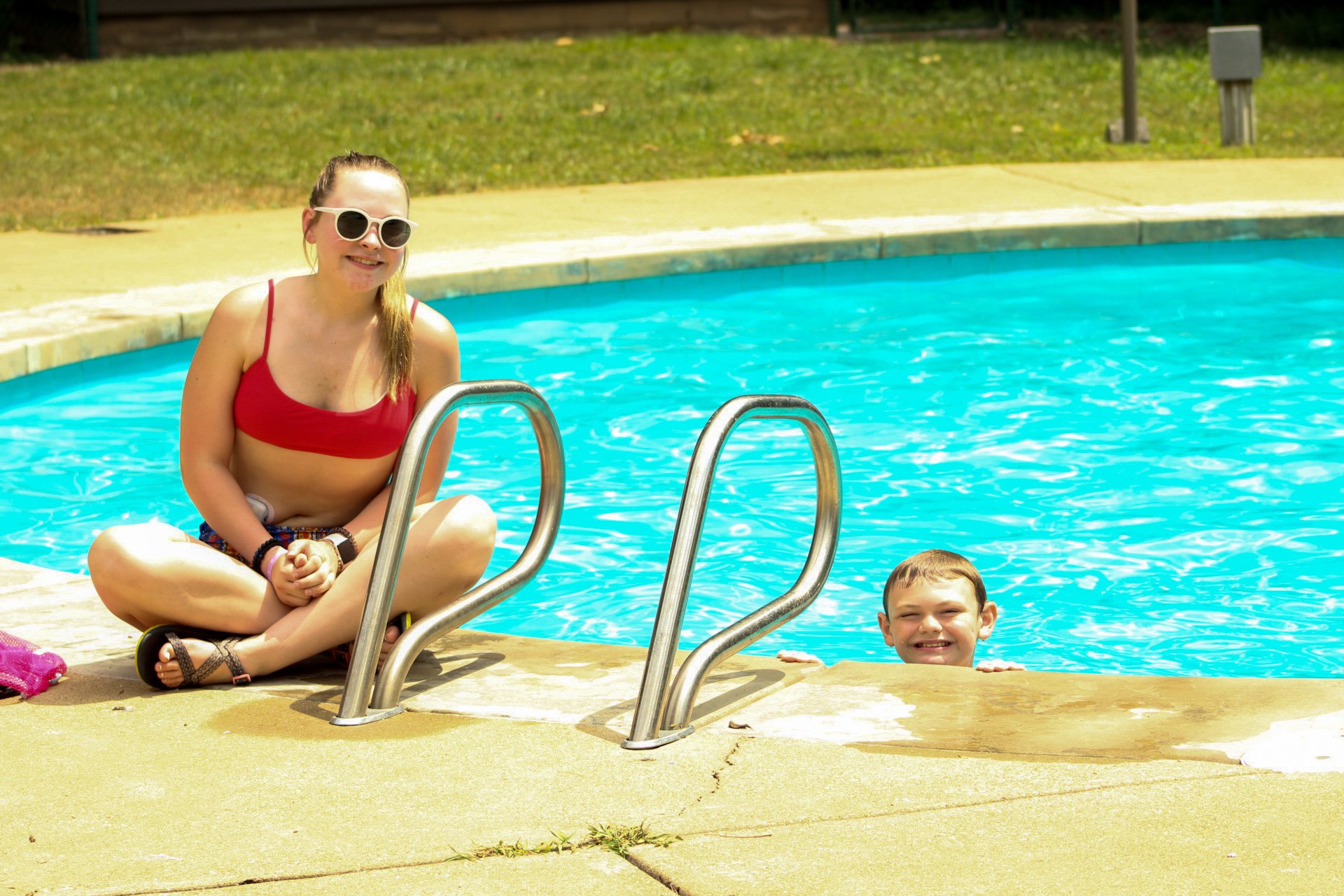 An older girl wearing a red bathing suit is sitting on the edge of the pool near the ladder. A boy is in the pool; both are smiling towards the camera