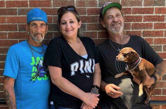 Three people standing against a brick wall, with one holding a small brown dog. The person on the left wears a blue shirt and blue cap, the person in the middle wears a black shirt with 'PINK' on it, and the person on the right wears a dark t-shirt and baseball cap. The dog has a collar and is being held gently.