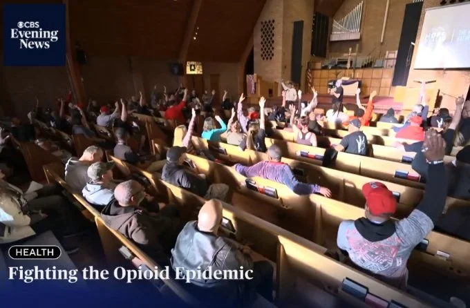 Audience in a church or auditorium with many people raising their hands during a presentation on fighting the opioid epidemic, as shown in CBS Evening News.