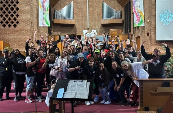 A large group of diverse people at The Hope Gathering recovery meeting. Some are making peace signs, others are smiling and raising their hands, with banners and an organ pipe visible in the background.