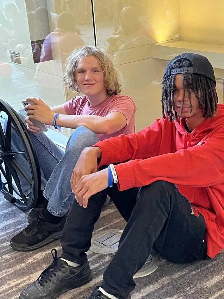 Two young men sitting on the floor next to a glass wall. The man on the left has curly blond hair, is wearing a pink T-shirt, and is holding a smartphone. The man on the right has dreadlocks, is wearing a black cap backward, a red hoodie, and black pants. They are inside a modern building with a yellow-lit wall behind them.