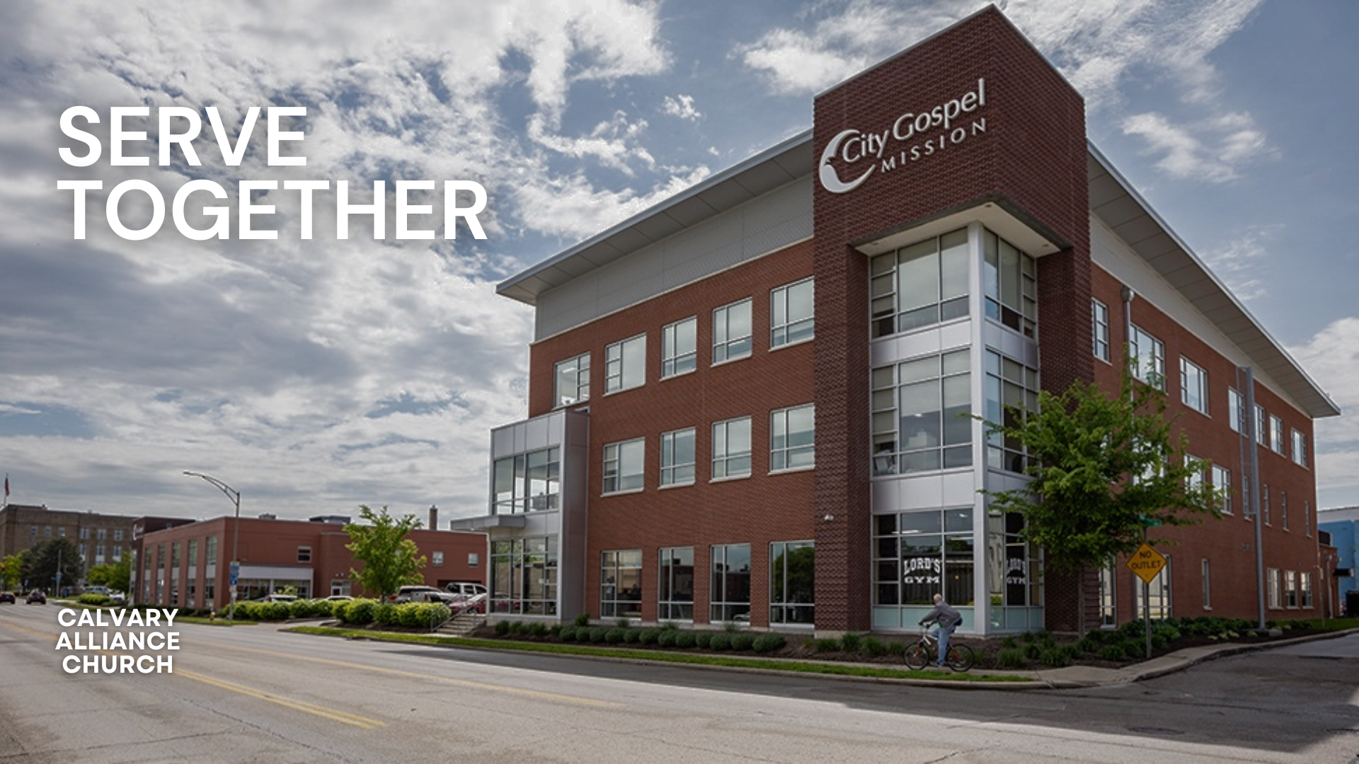 A multi-story brick and glass building with the sign 'City Gospel Mission' on the corner, situated on a city street with a cyclist and green trees in front, and the text 'Serve Together' and 'Calvary Alliance Church' overlayed.