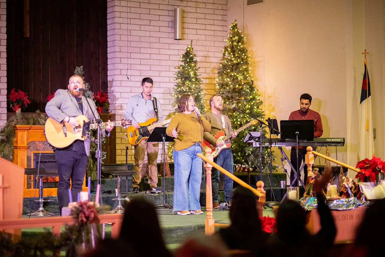 A musical group performing on stage with Christmas trees and poinsettias in the background, in what appears to be a church or community hall decorated for the holiday season.