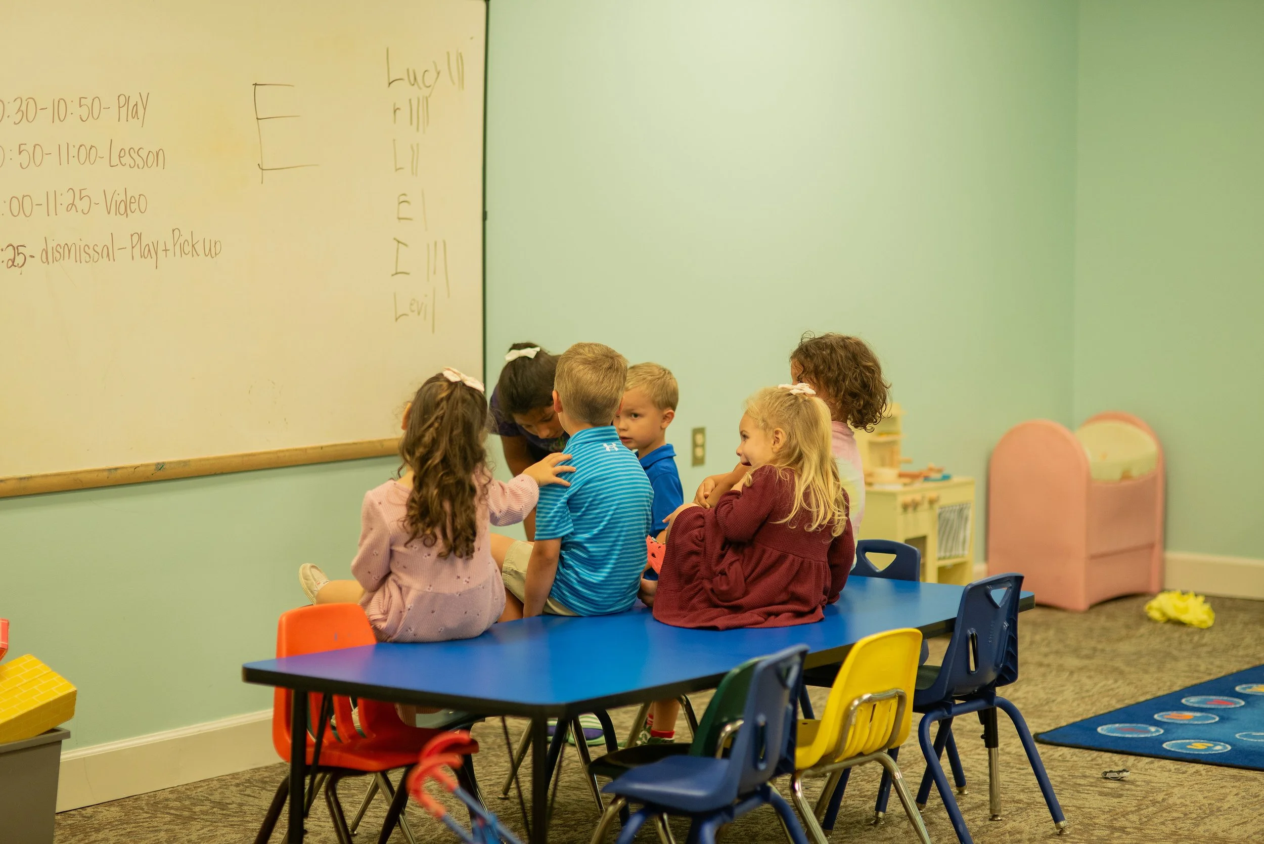 Preschool classroom with teacher and children engaged in learning activity, colorful tables and chairs, educational materials visible.