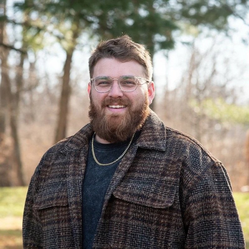 A smiling man with glasses and a beard outdoors in a forested area.