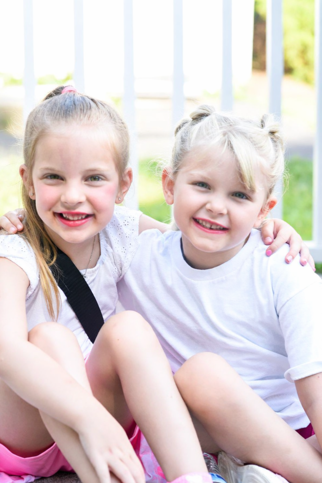 Two young girls smiling and sitting outdoors, hugging each other, on a sunny day with a blurred background of green grass and a white railing.