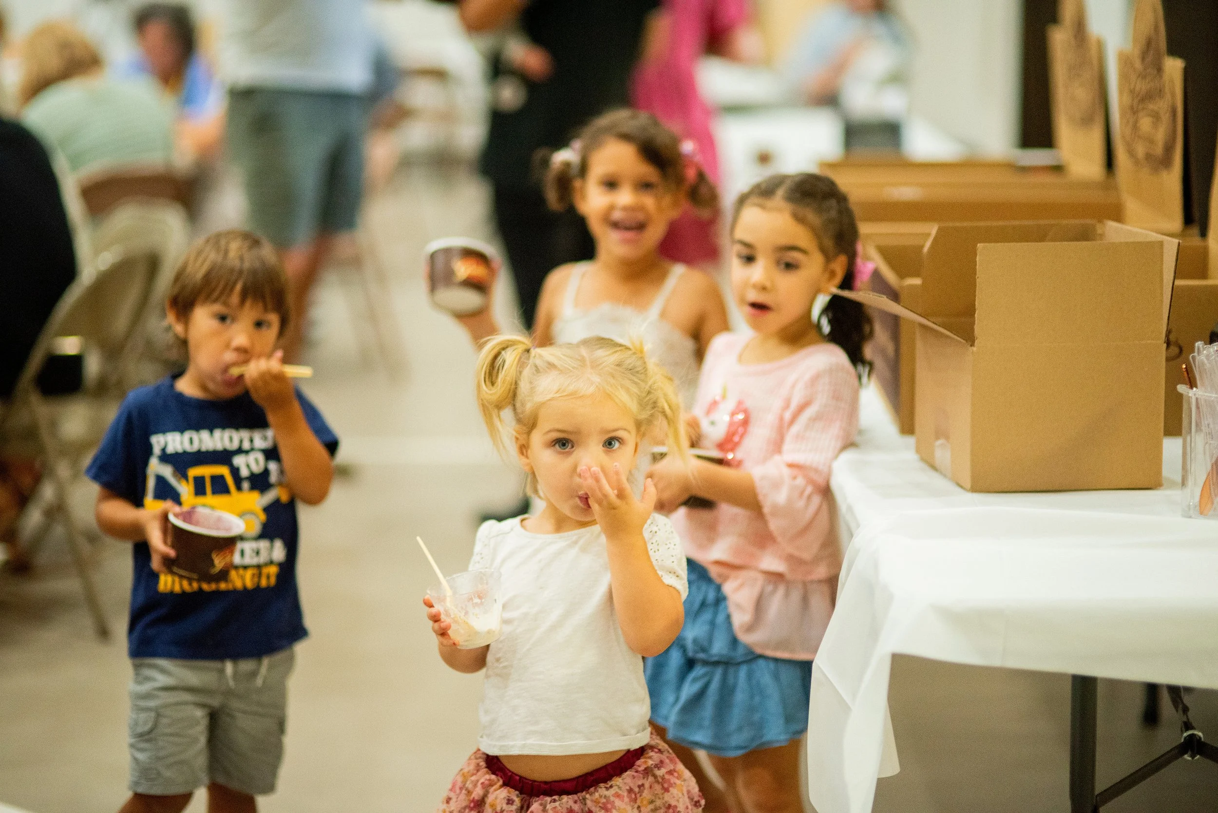 Group of five children standing in line at what appears to be a party or event, with tables, boxes, and people in the background.