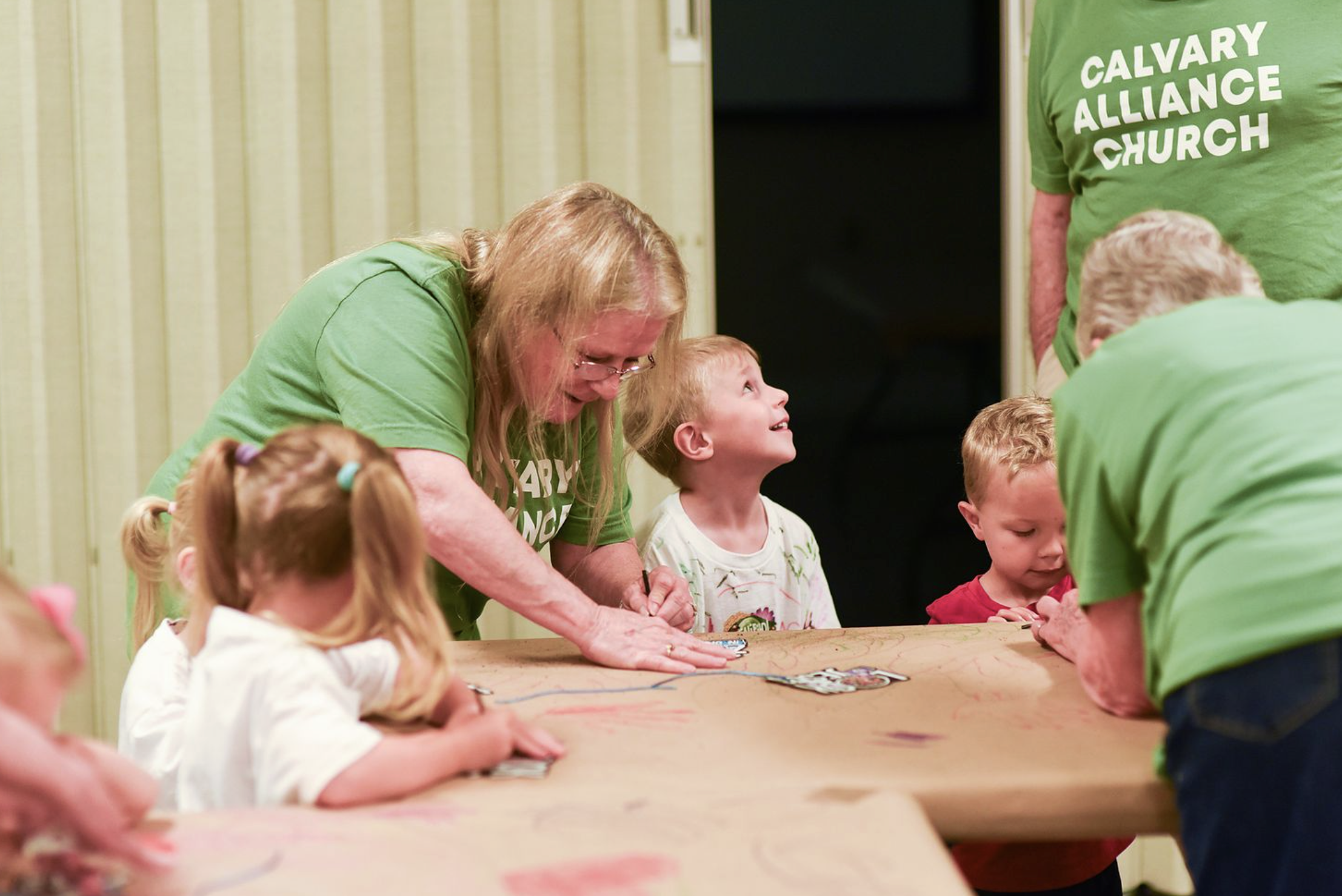 A group of children and adults in green Calvary Alliance Church shirts gathered around a table, engaging in an activity indoors.