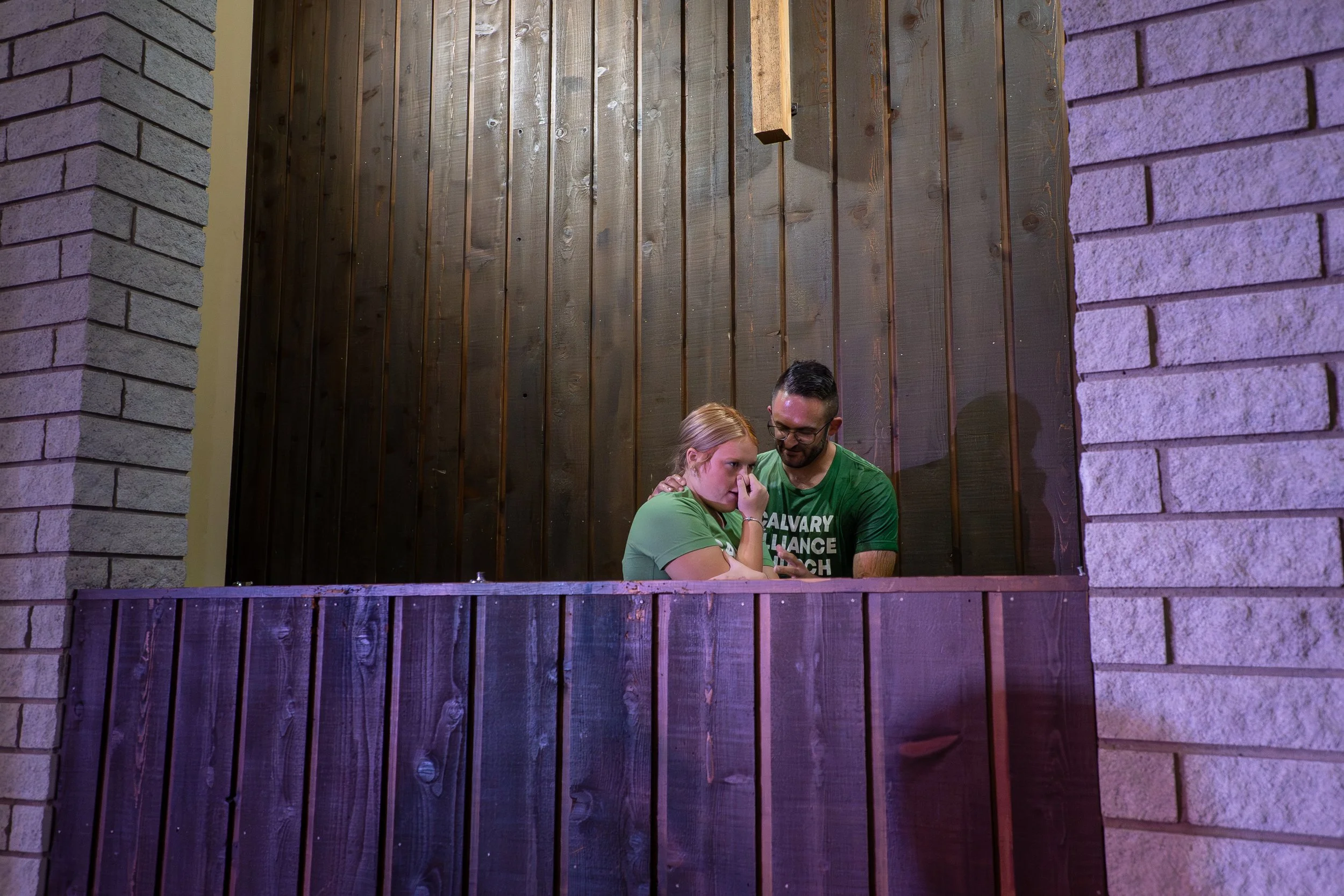 A man and a young girl, both wearing green shirts, stand on a balcony with a wooden wall behind them. The girl looks distressed, holding her nose, while the man comforts her, leaning in close and smiling.