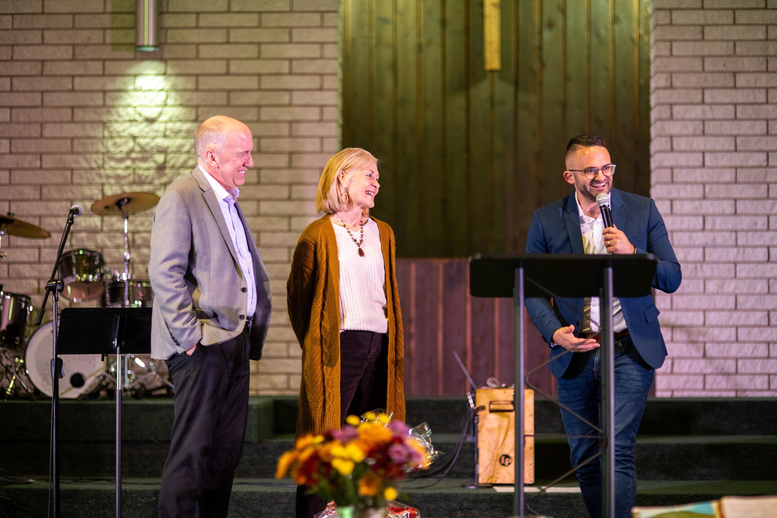 Person speaking on stage in a church setting, with microphone and lectern, against a backdrop of brick walls and religious symbols.