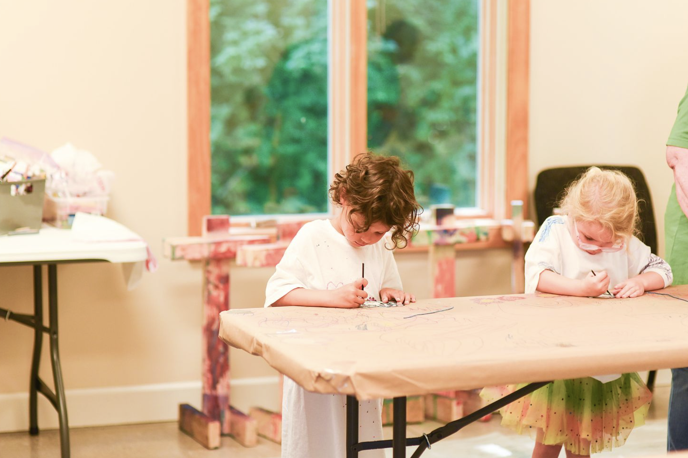 Two young girls with art supplies drawing on a large piece of paper at a table in a bright indoor space with a large window showing green trees outside.