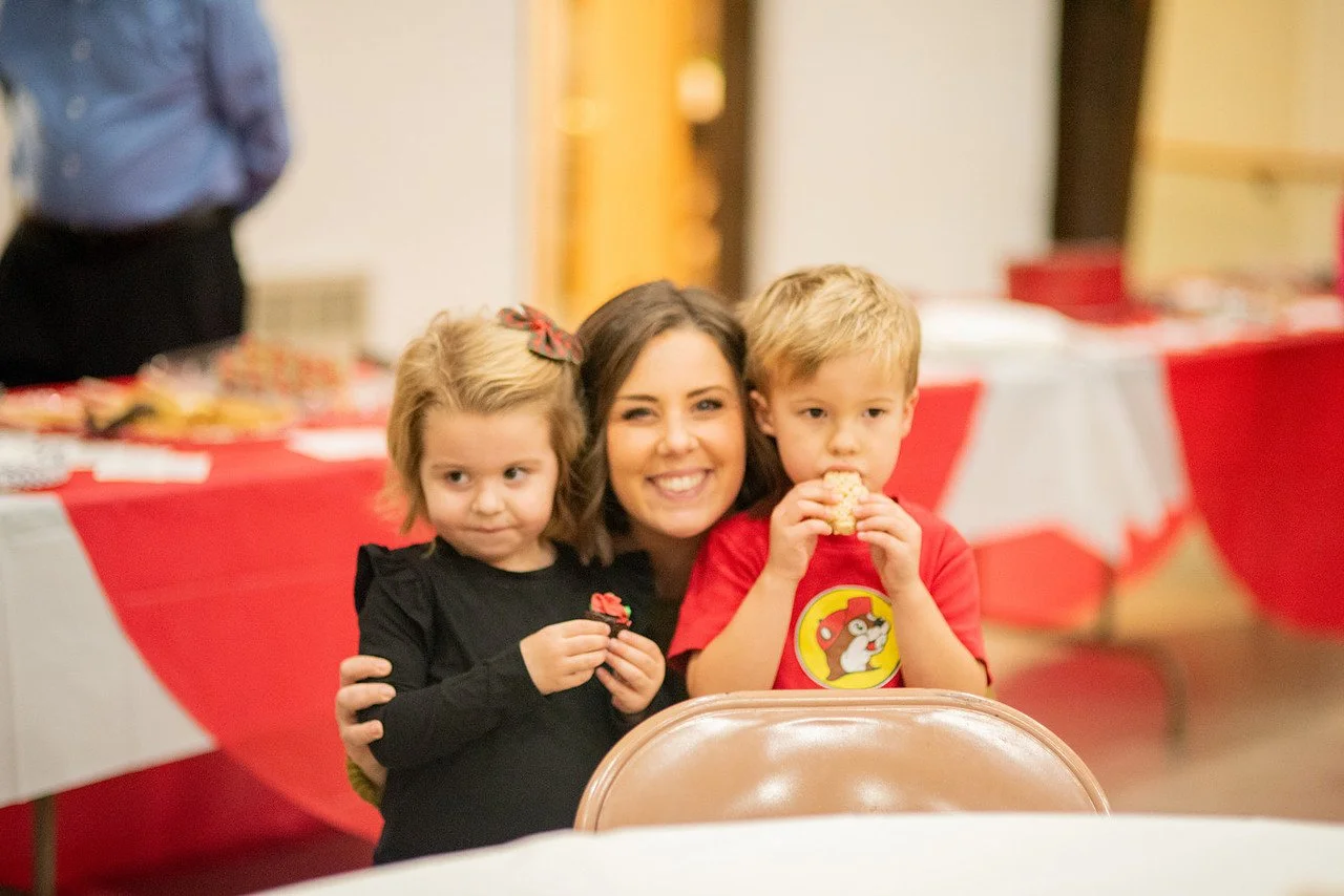 A woman with two children at a table during a party, with red and white decorations, enjoying snacks.