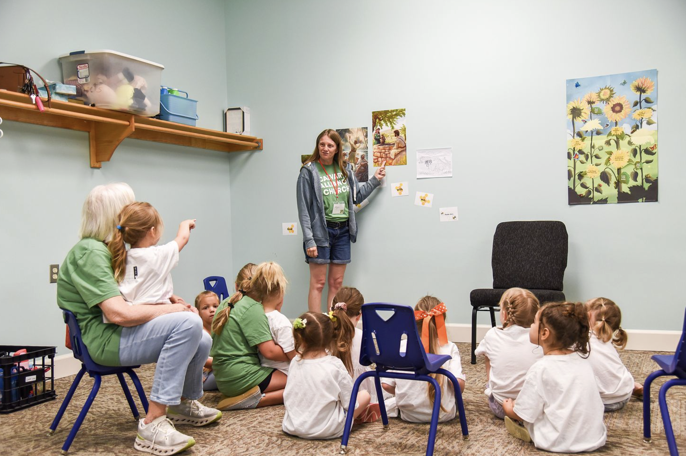 A woman teaching a group of young children in a classroom, with children sitting on the floor and some sitting on small blue chairs, as she points to pictures on the wall, including a large sunflower poster.