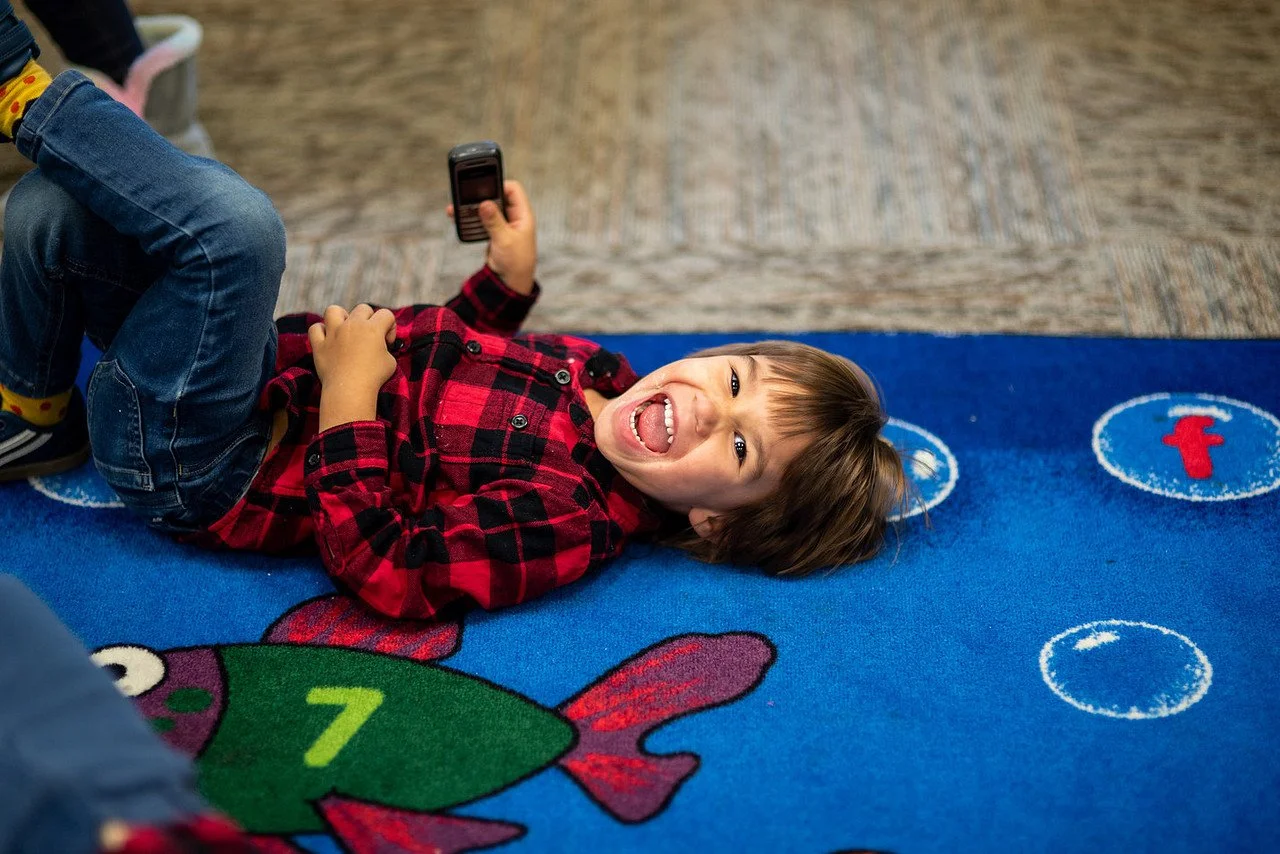 A young boy lying on a colorful carpet, smiling and holding a phone, wearing a red and black plaid shirt and blue jeans.