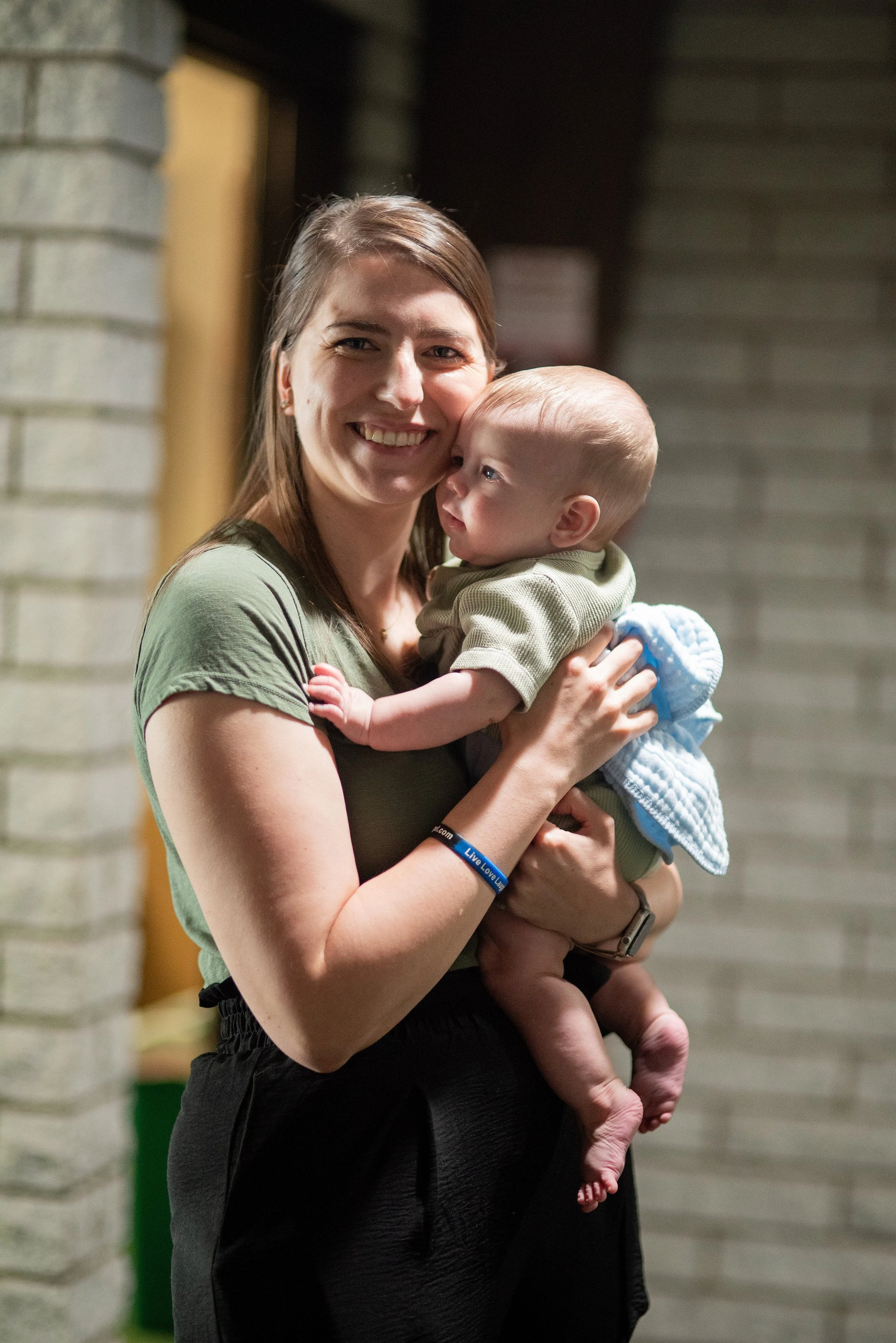 Smiling woman holding a baby with a beige shirt and a blue blanket in front of a brick wall.