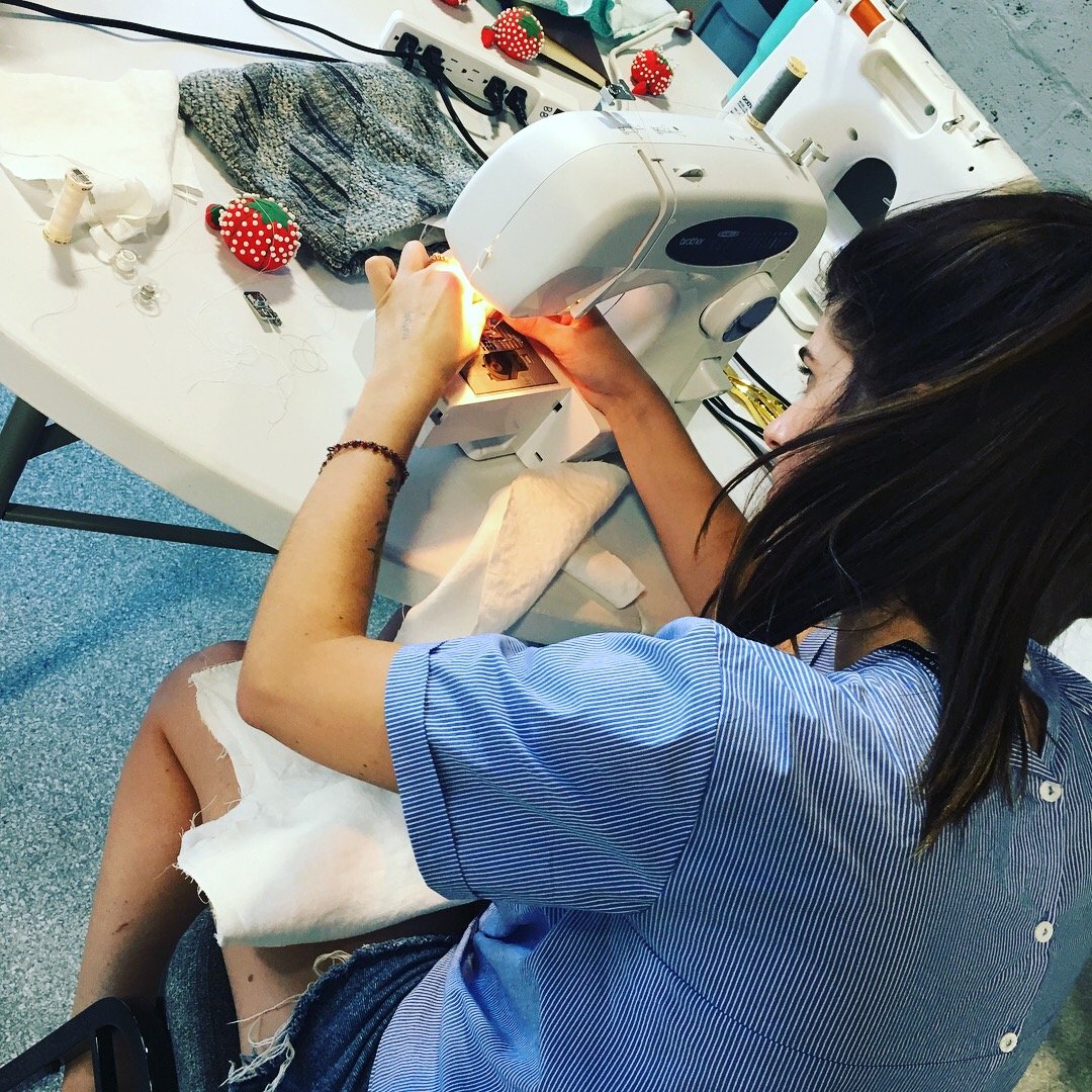 A woman sewing fabric with a sewing machine at a workspace. The table has sewing supplies, fabric, and stuffed toys nearby.
