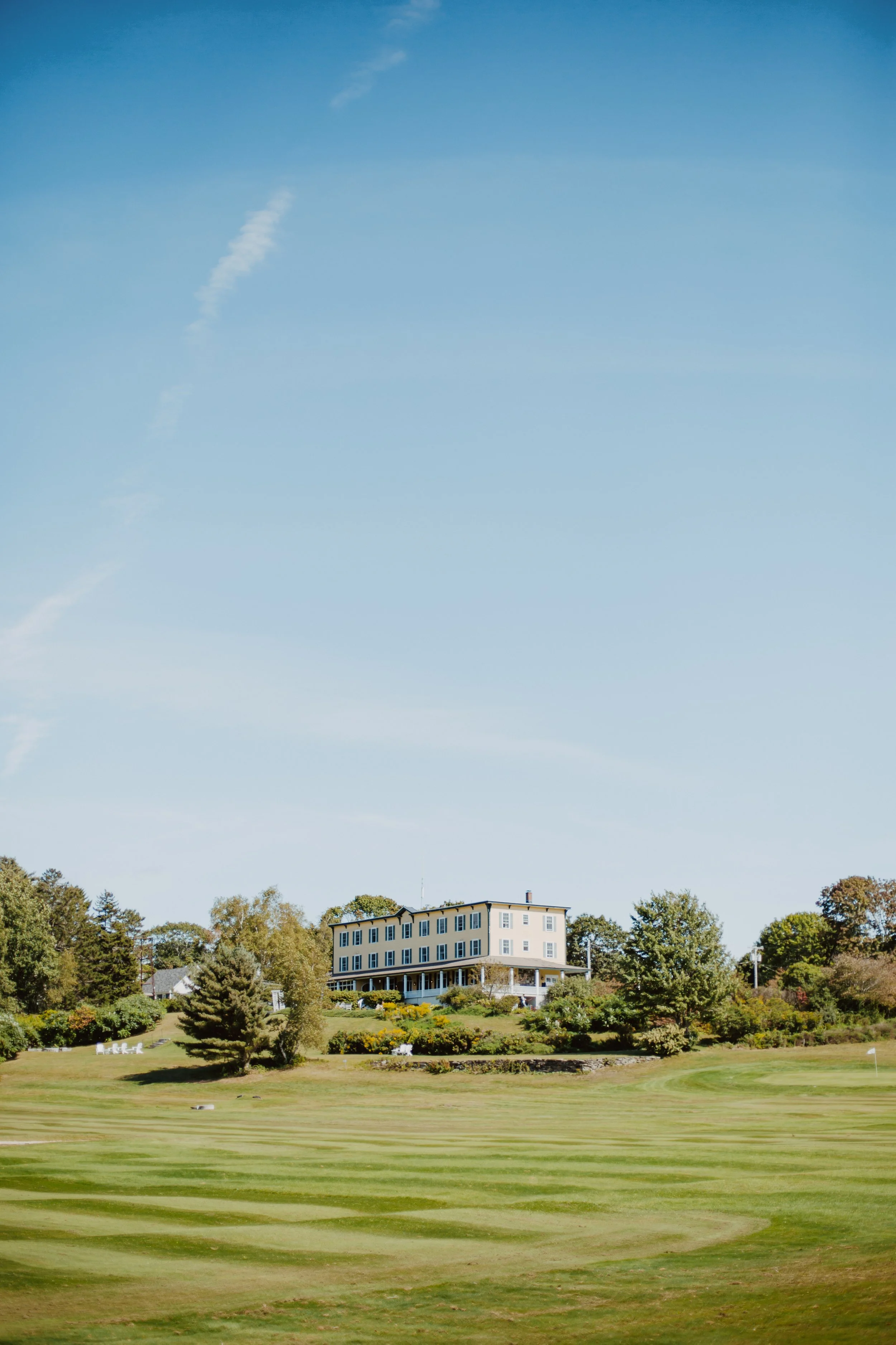 A large white building on a hill overlooking a green golf course with trees around.