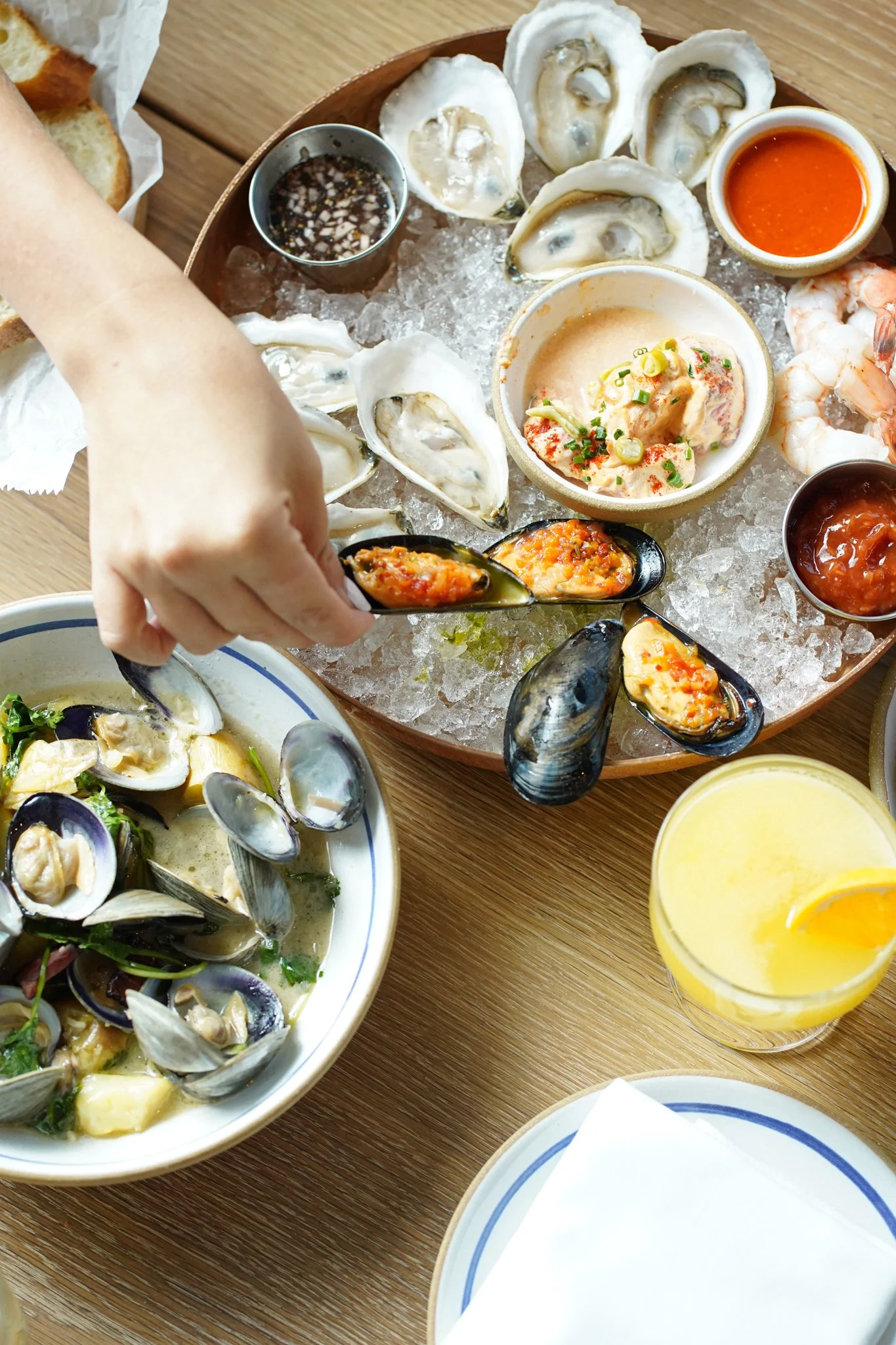 Seafood platter with oysters, clams, shrimp, and dipping sauces on a bed of ice, with a person's hand picking up stuffed mussels, and lemon drinks on a wooden table.