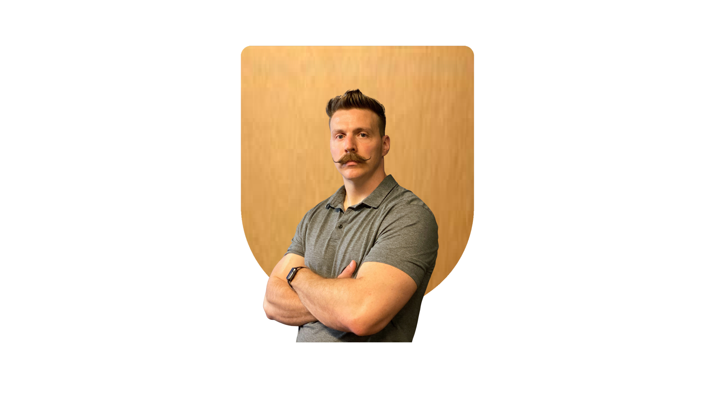 A young man with styled hair and a curled moustache, wearing a grey polo shirt, poses with his arms crossed against a wooden background.