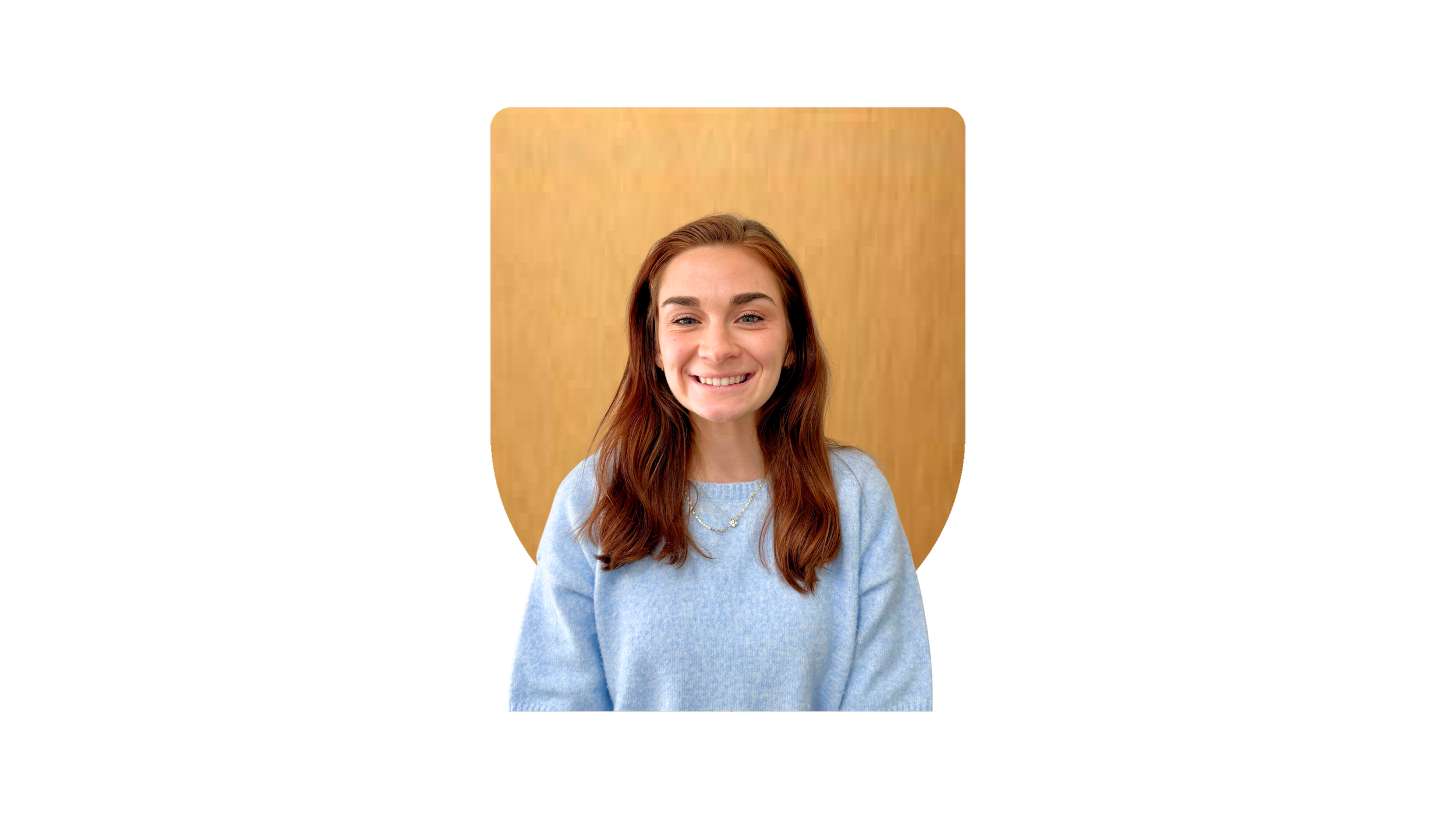 A young woman with long brown hair, smiling, wearing a light blue sweater and a necklace, sitting in front of a wooden background.